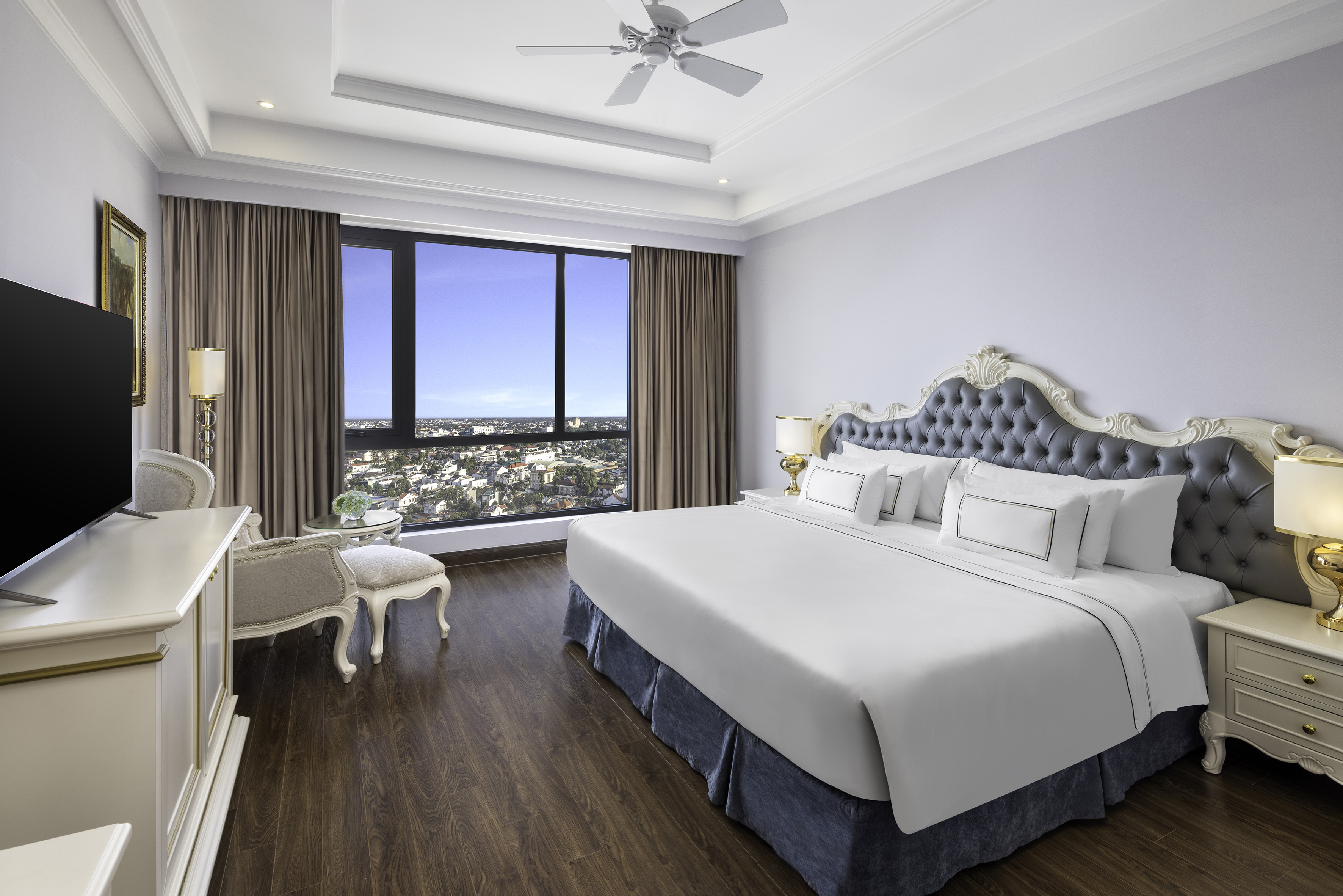 Luxurious hotel room with large bed, grey tufted headboard, and panoramic city view.