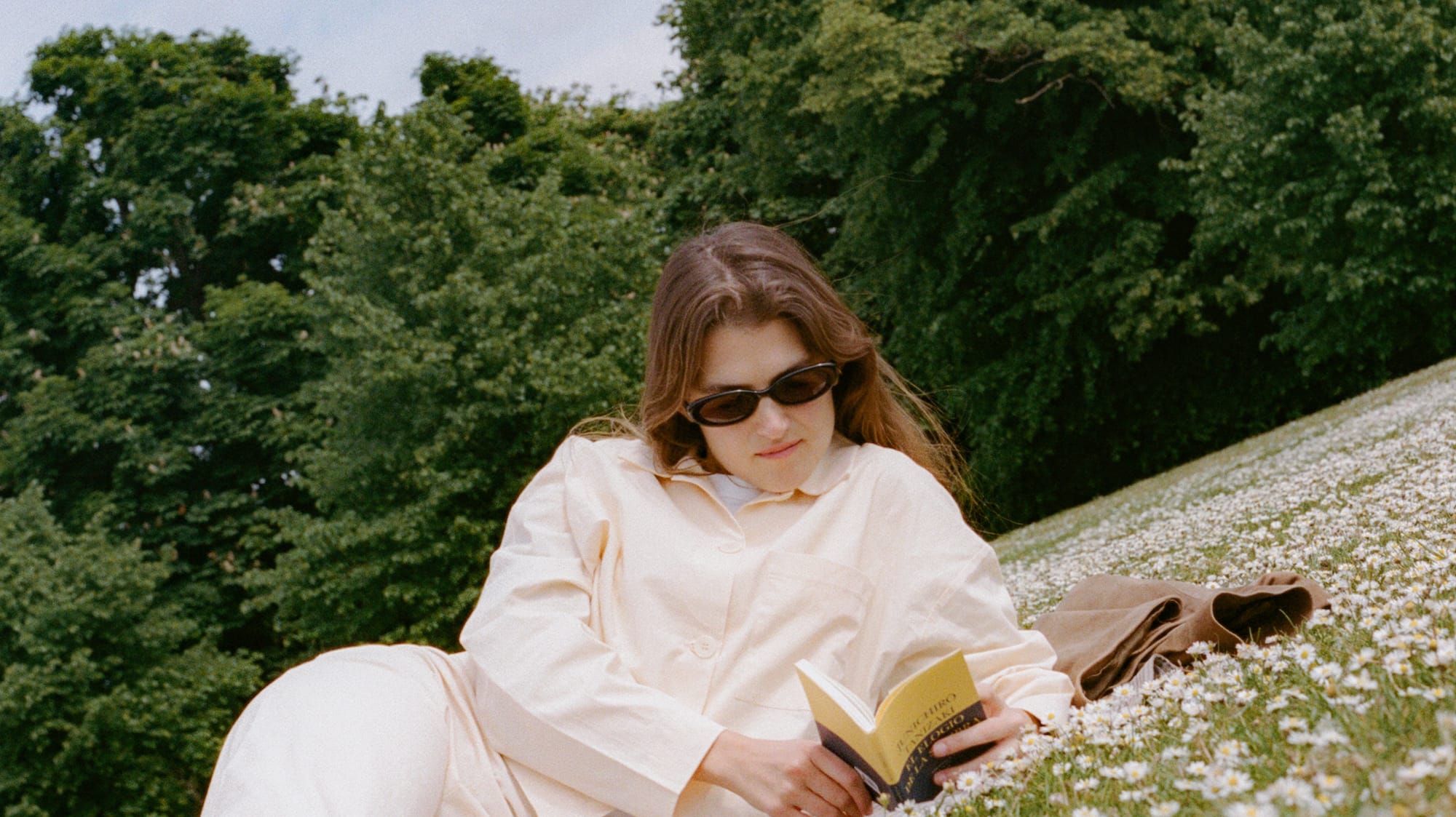 Woman in sunglasses reading book, relaxing on a daisy-covered grassy hill.