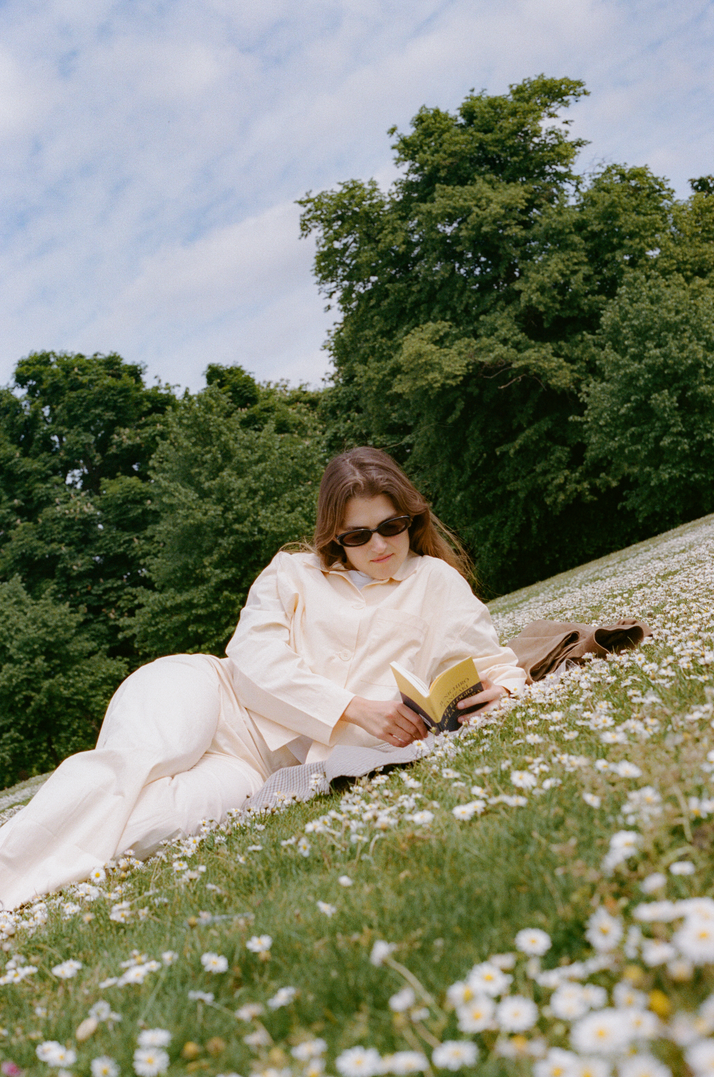 Woman in sunglasses reading book, relaxing on a daisy-covered grassy hill.