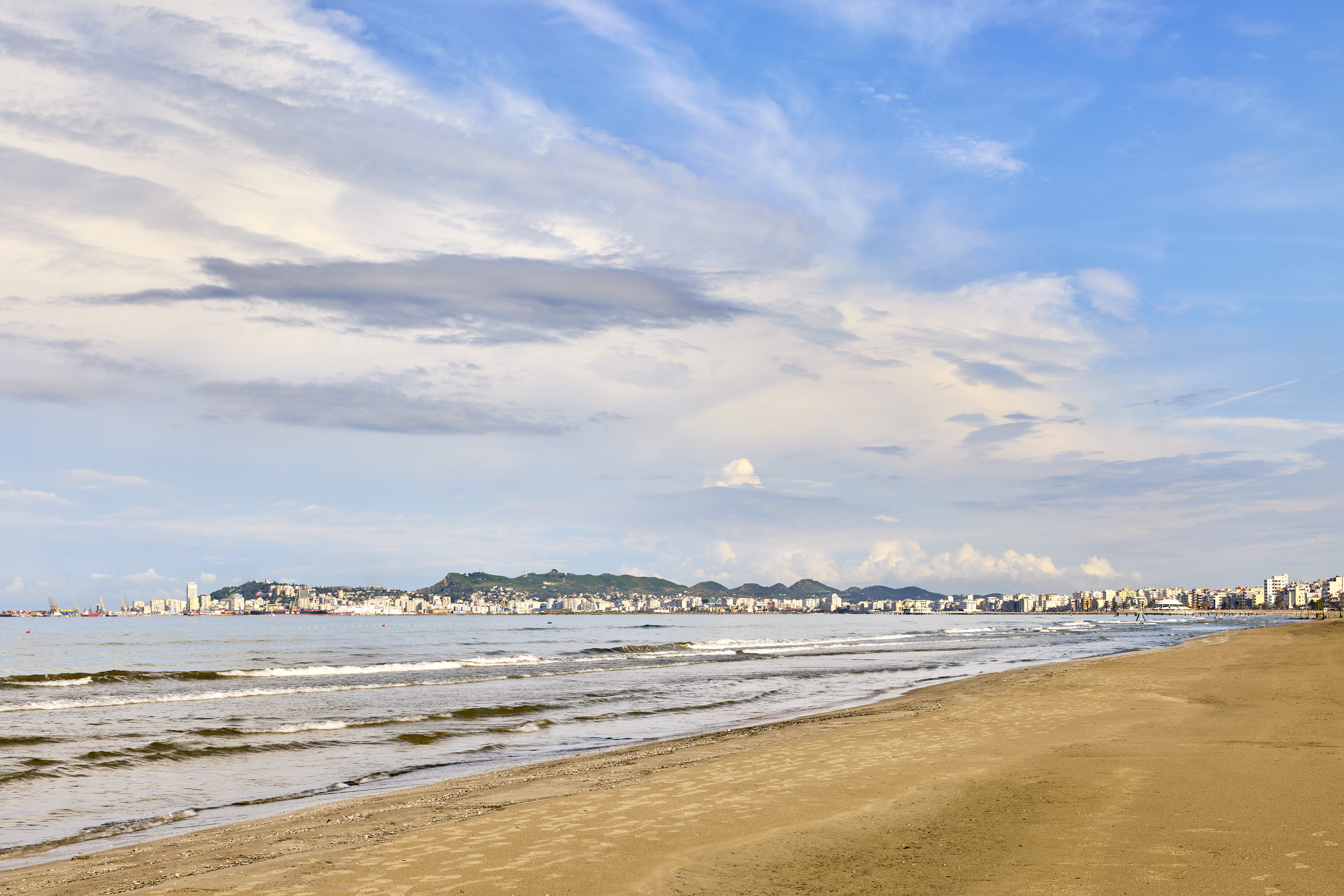 Coastal city view from sandy beach with gentle ocean waves and clouds.