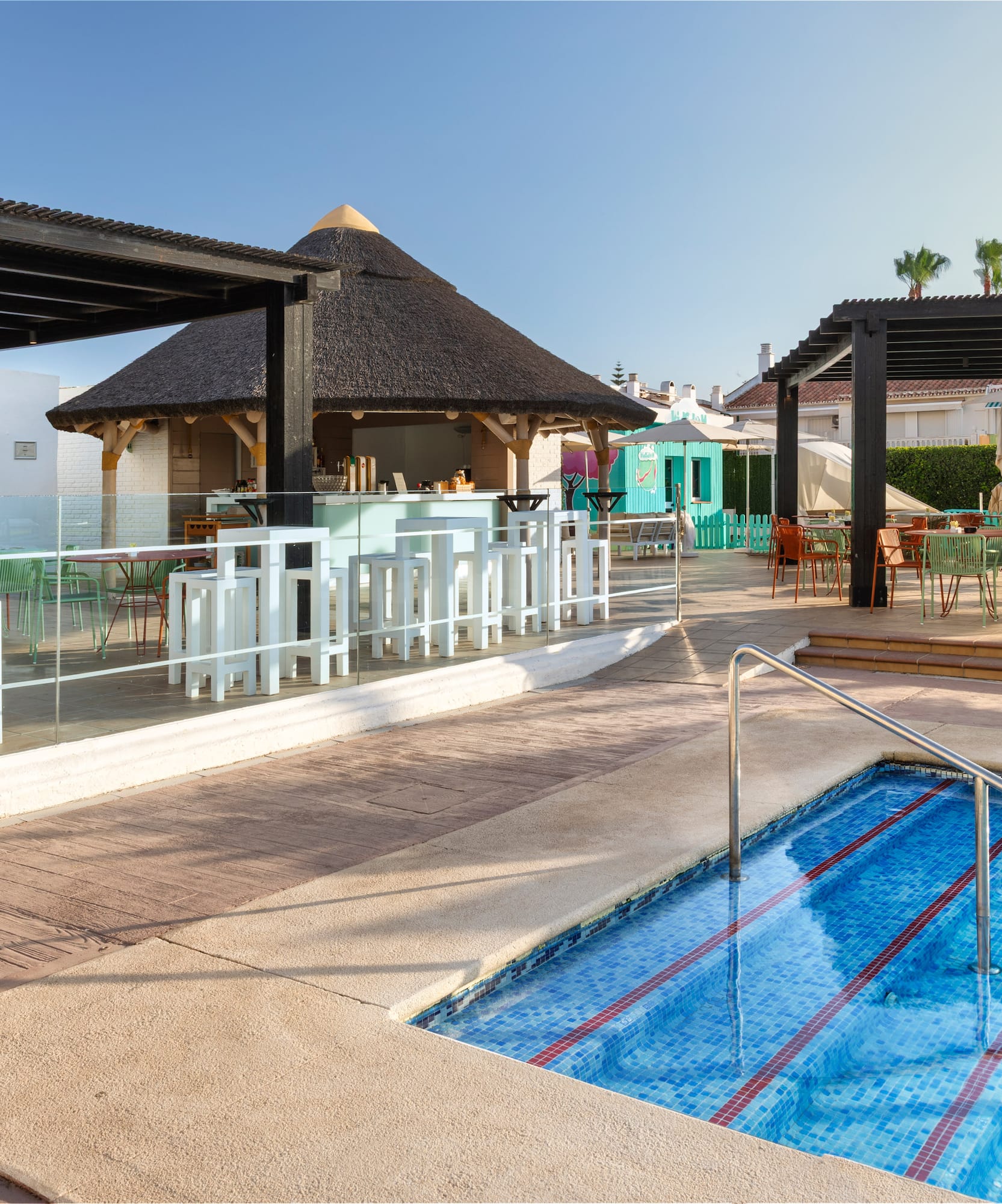 Sunny resort pool with thatched bar and shaded outdoor dining areas.