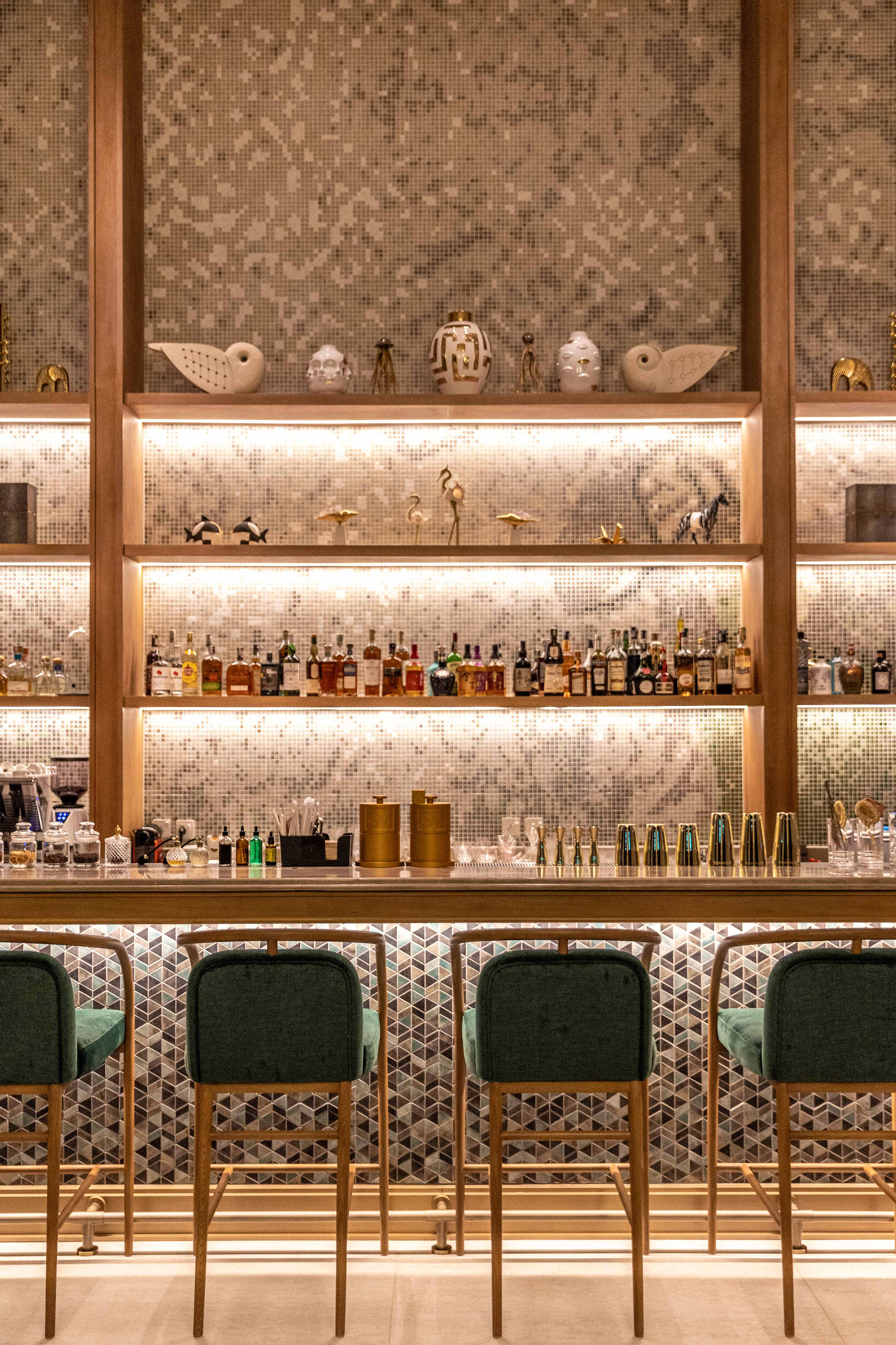 Illuminated bar with liquor bottles on mosaic shelves, green velvet bar stools.