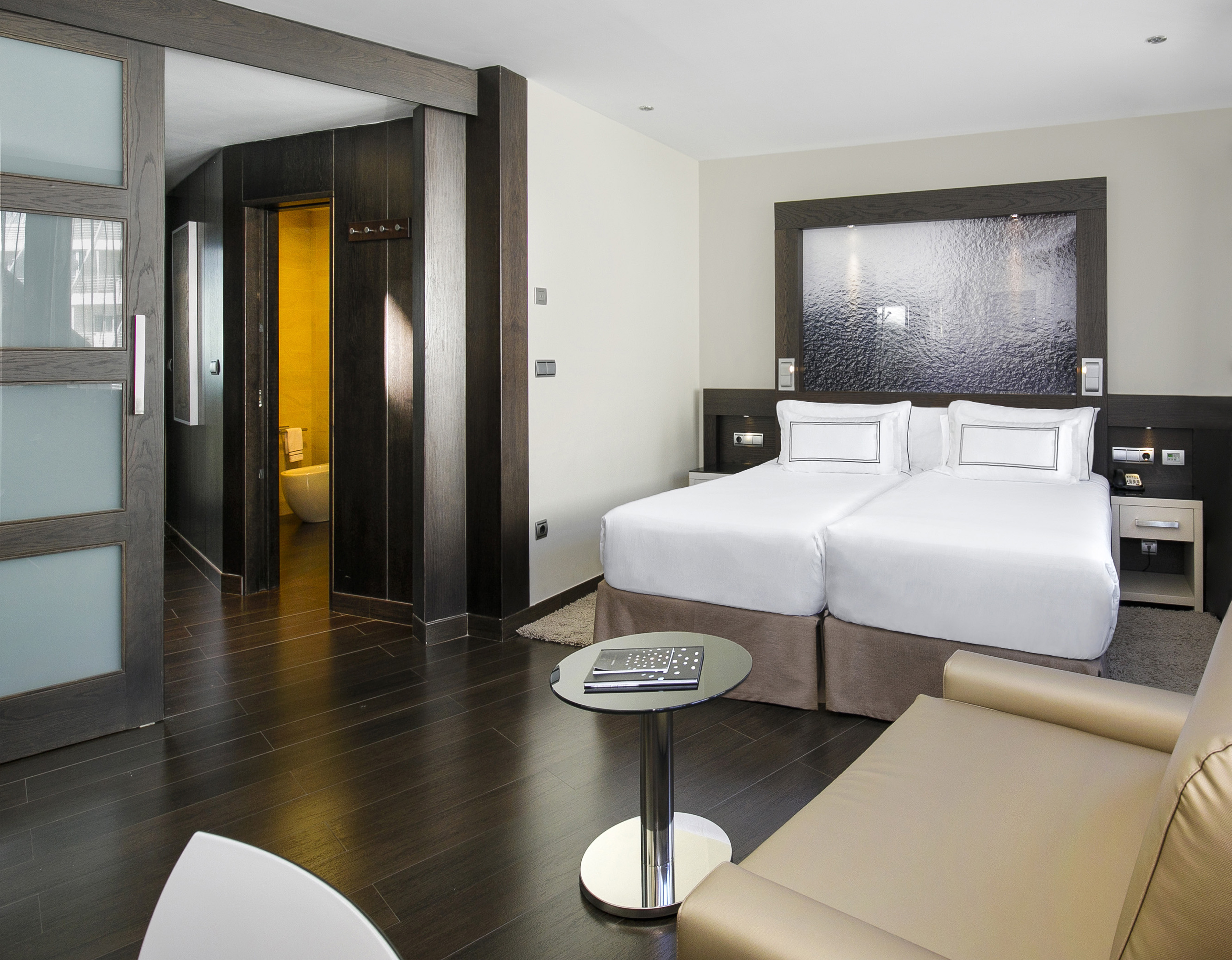 Modern hotel room with twin beds, dark wood interior, and en-suite bathroom.