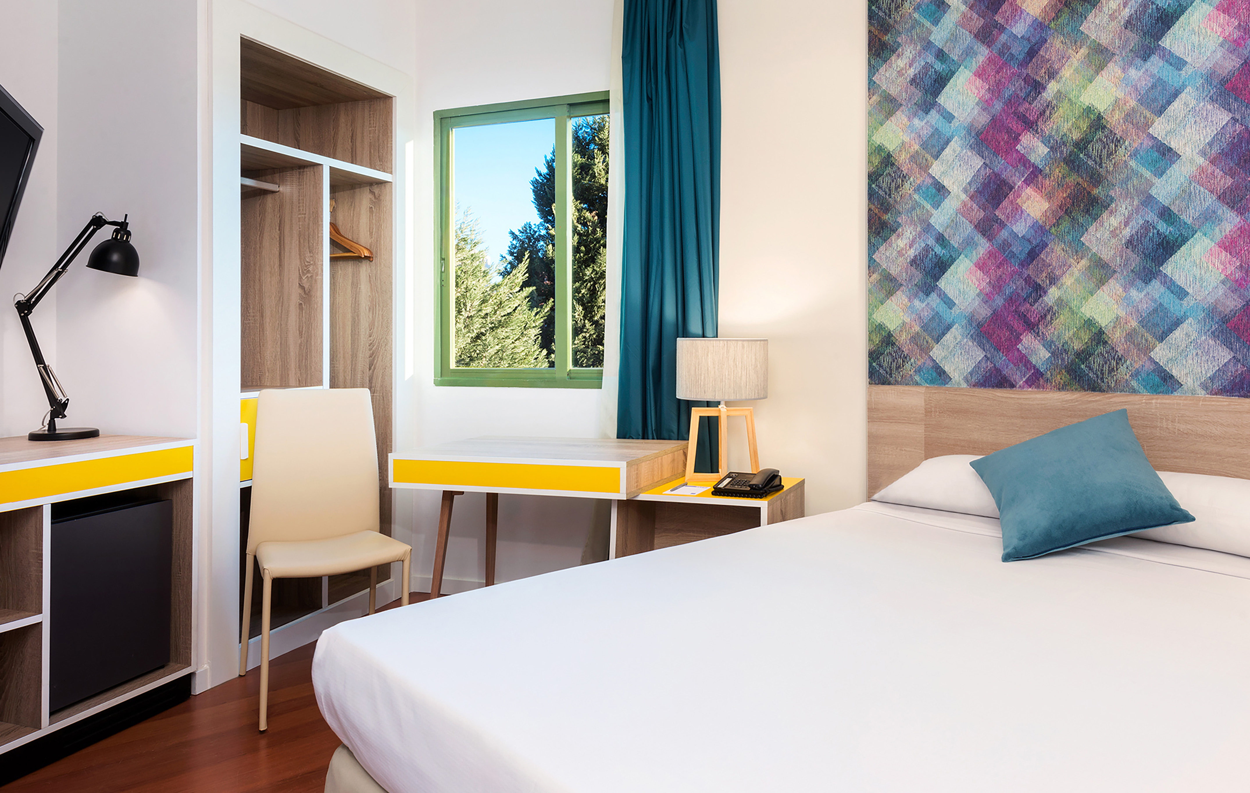 Modern hotel room with colorful geometric headboard, desk, and window.