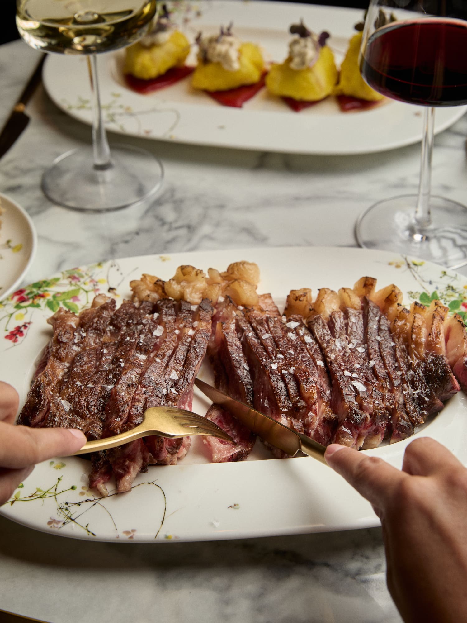 Sliced steak with sea salt, gold cutlery, wine, and small appetizers.