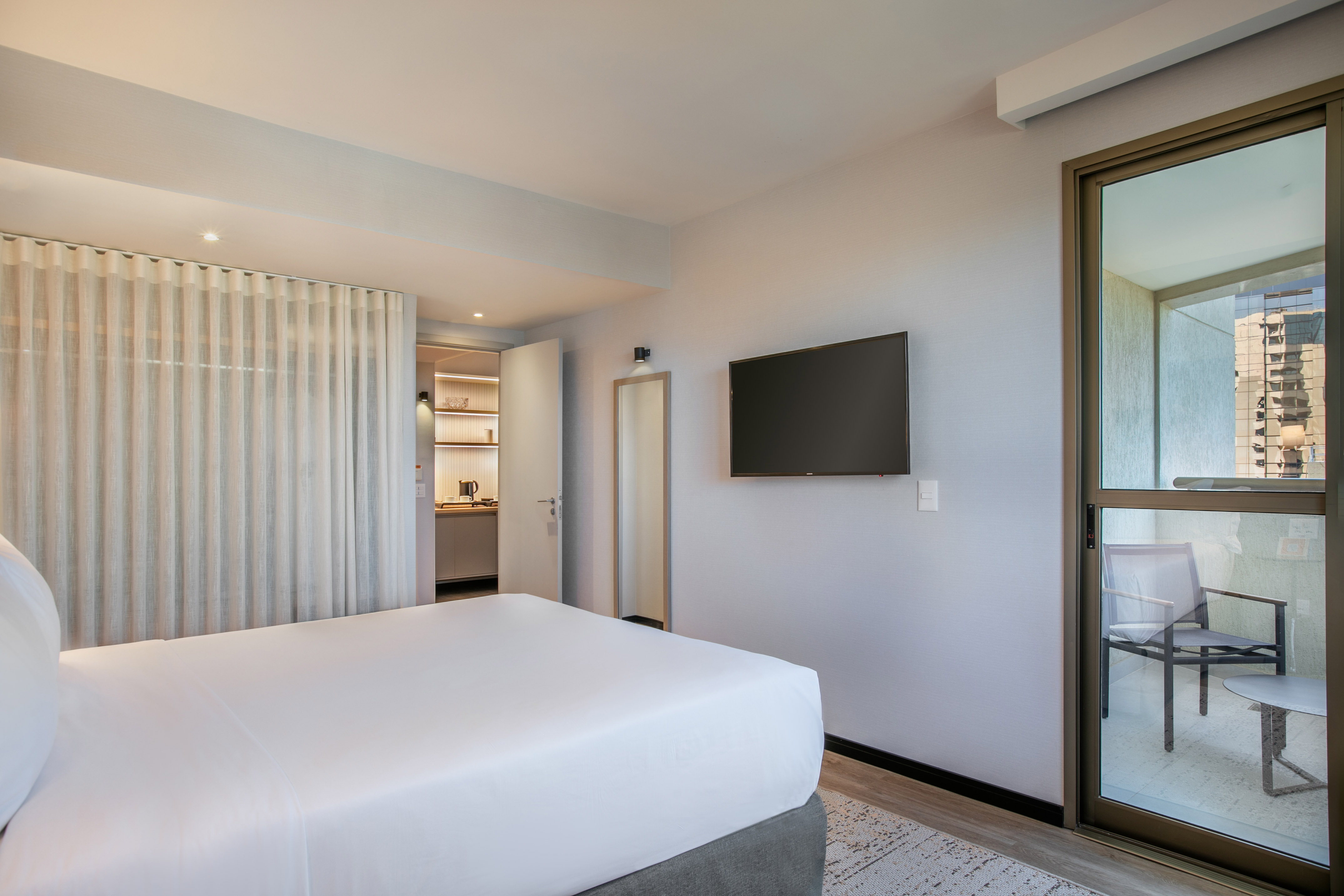 Modern hotel room with white bed, wall TV, compact kitchen, and balcony.