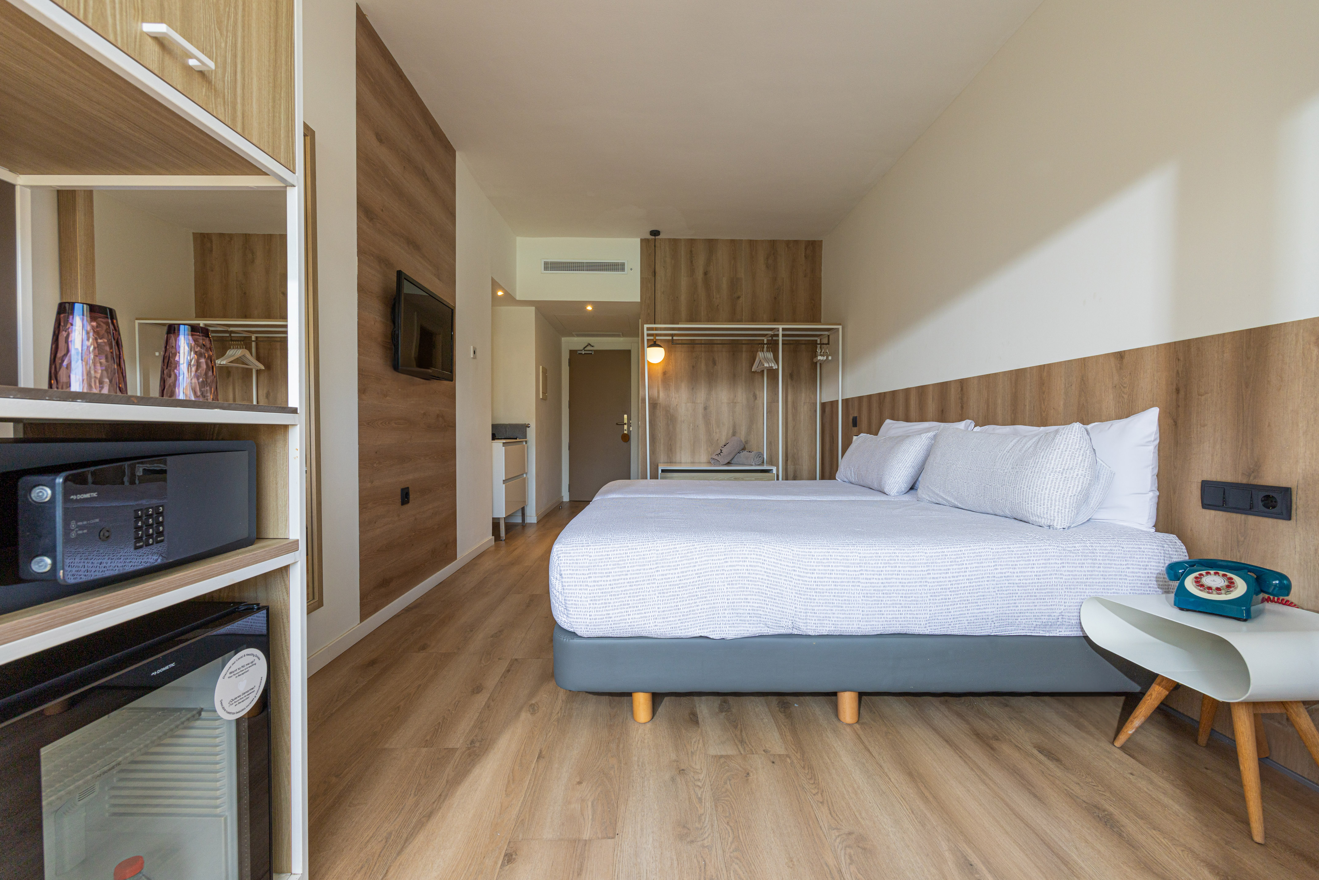 Modern hotel bedroom, large bed, wood decor, TV, mini-fridge, safe.