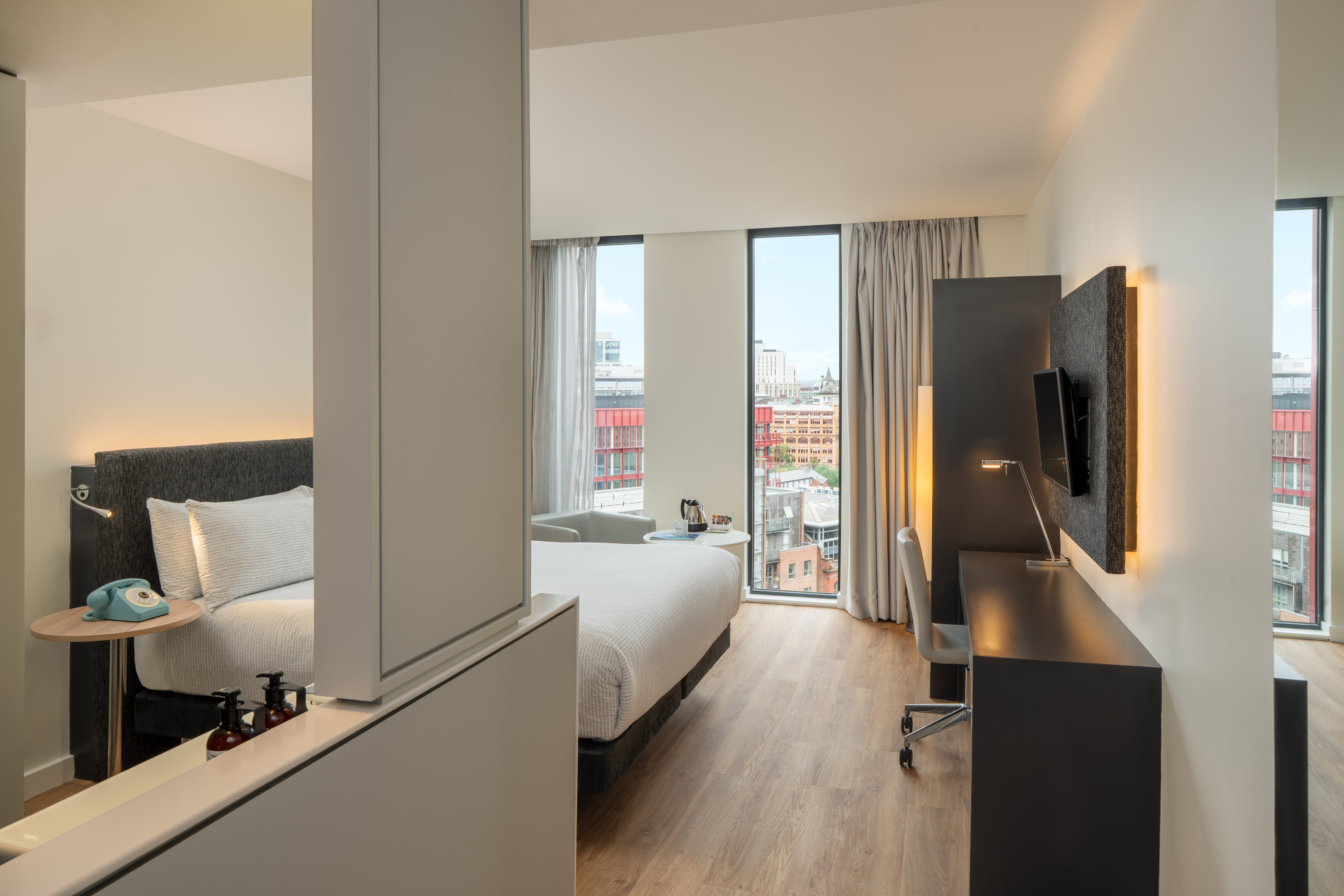 Modern hotel room with city view, bed, desk, and large windows.