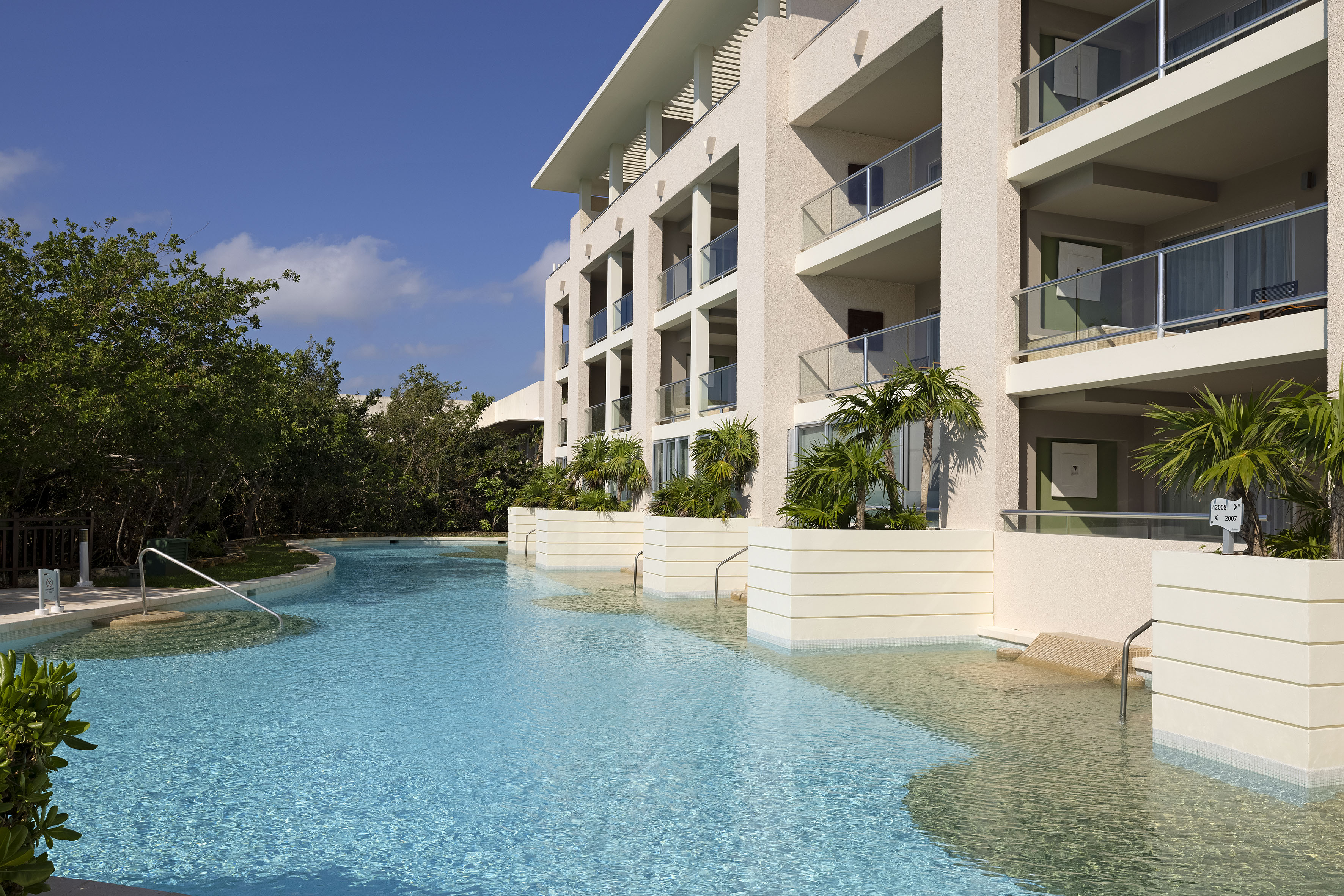 Tropical resort hotel with large swimming pool, palm trees, and modern balconies.