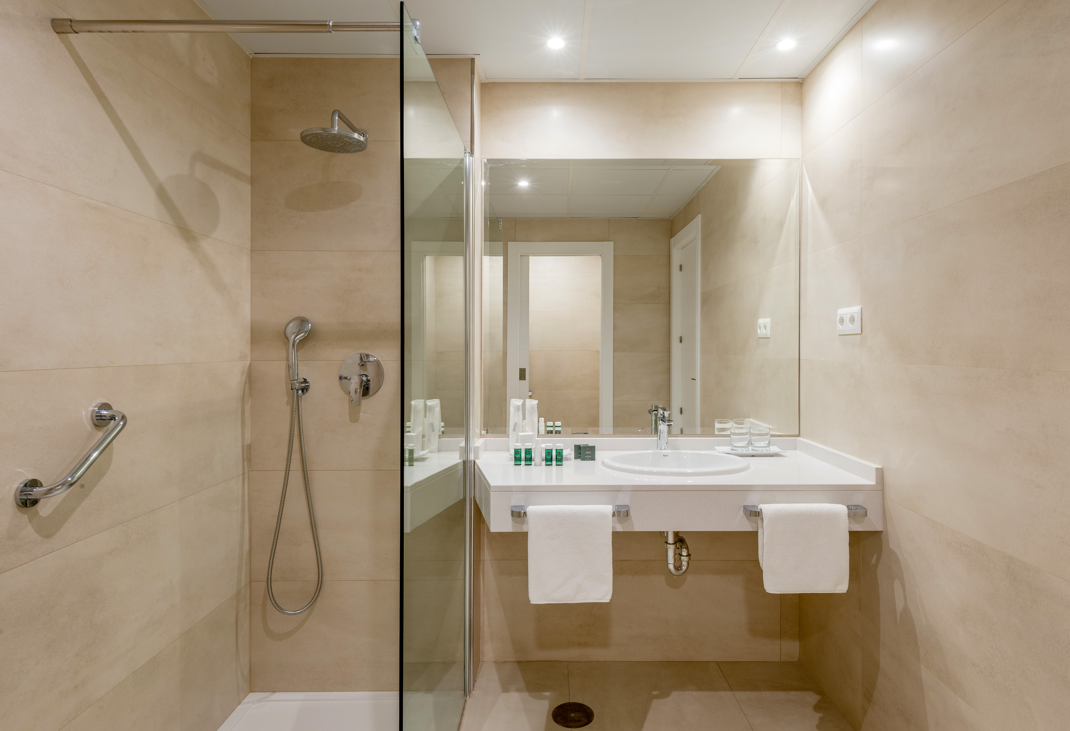 Modern hotel bathroom with walk-in shower, grab bar, vanity, sink, and beige tiles.