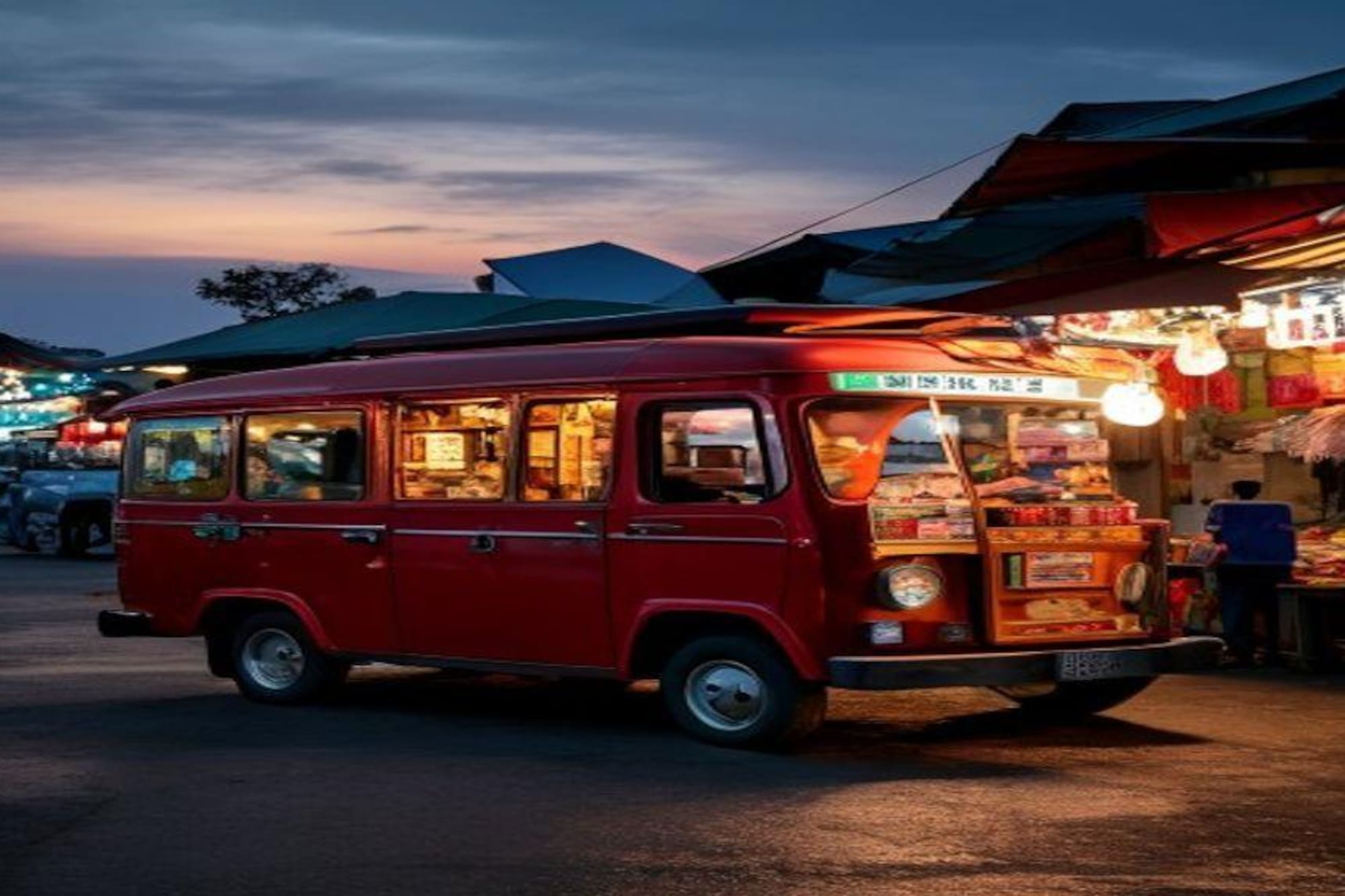 Red food truck brightly lit at a bustling night market under a twilight sky.
