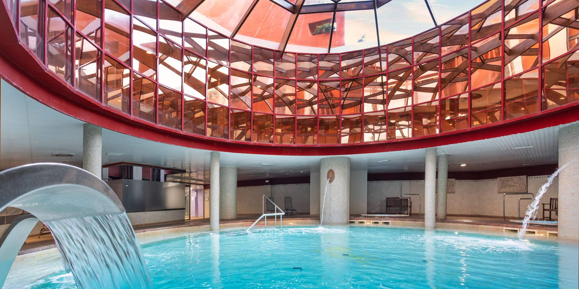 Indoor spa pool with waterfall and geometric red-framed glass dome roof.
