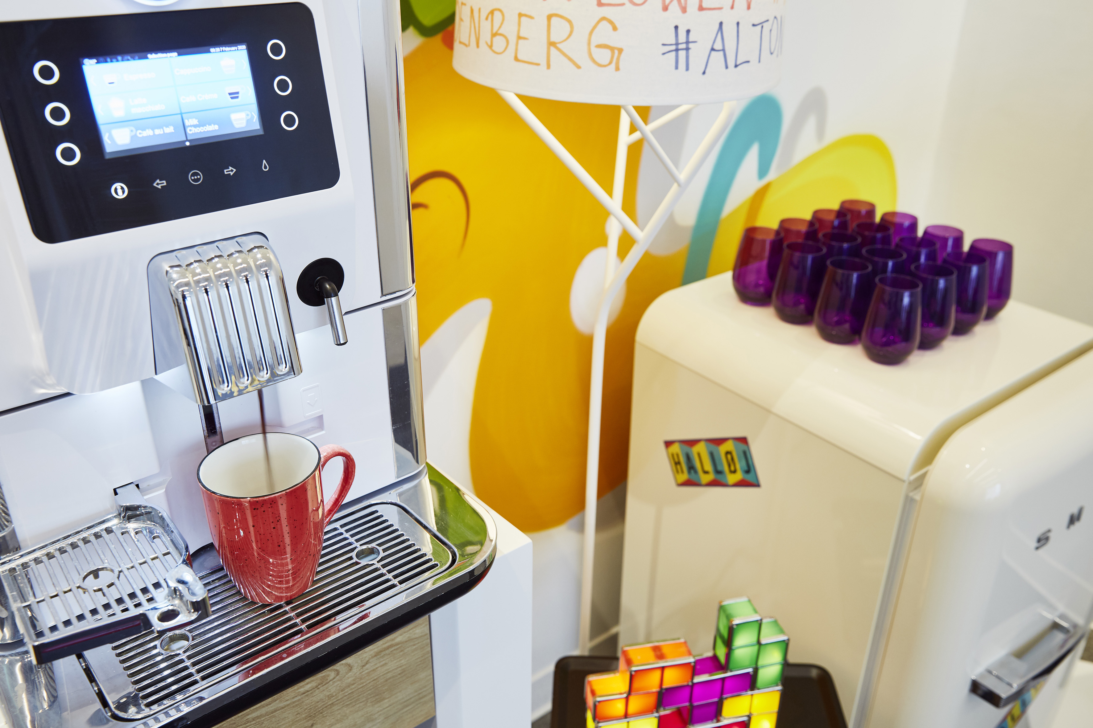 Espresso machine filling red mug, next to Smeg fridge and colorful wall decor.
