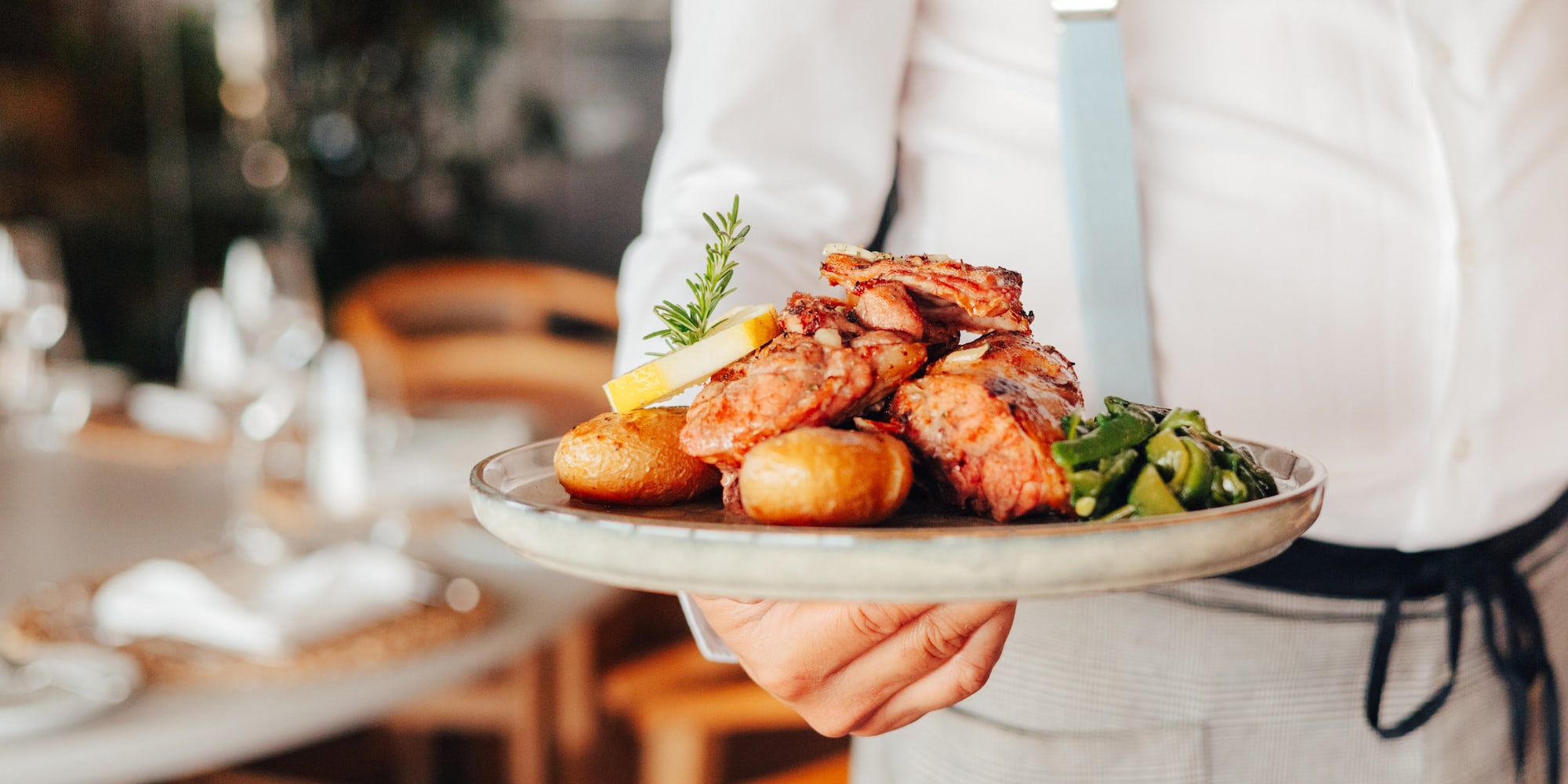 Waiter serving grilled meat, roasted potatoes, vegetables, lemon, and rosemary.
