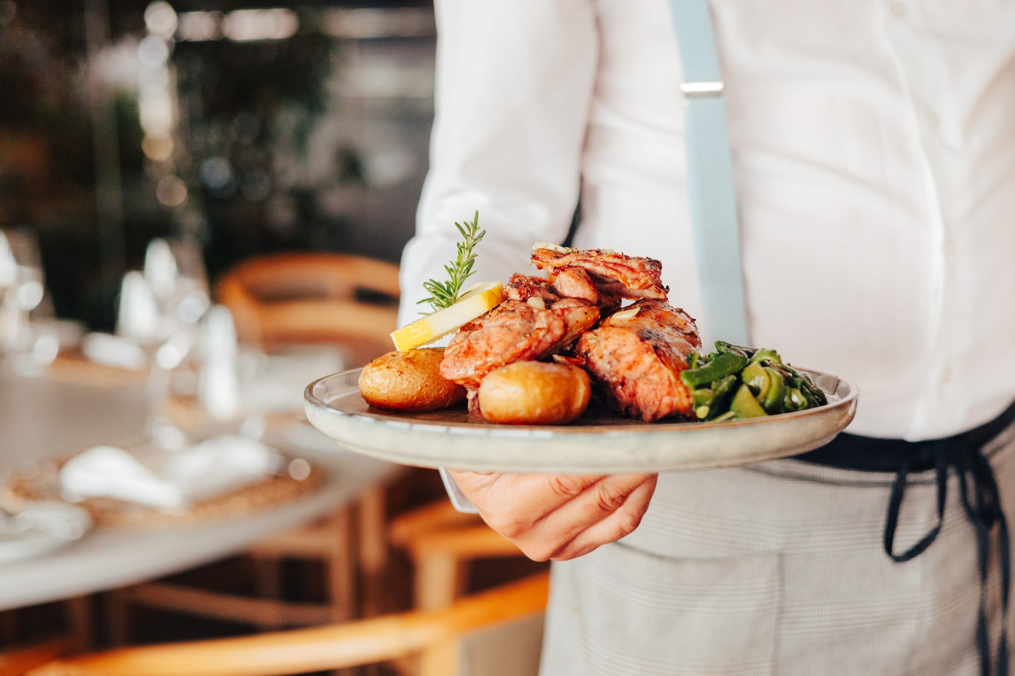 Waiter serving grilled meat, roasted potatoes, vegetables, lemon, and rosemary.