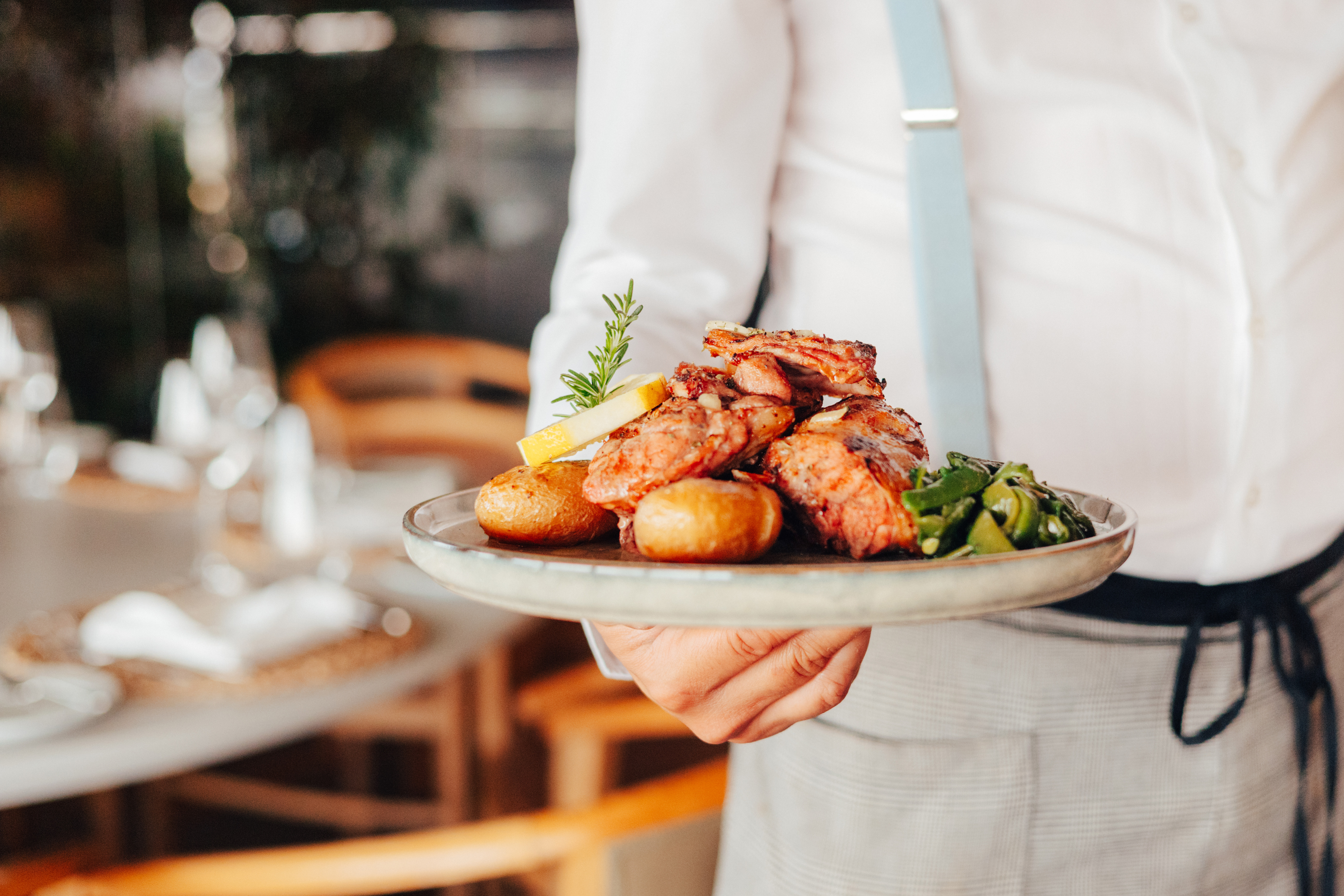 Waiter serving grilled meat, roasted potatoes, vegetables, lemon, and rosemary.