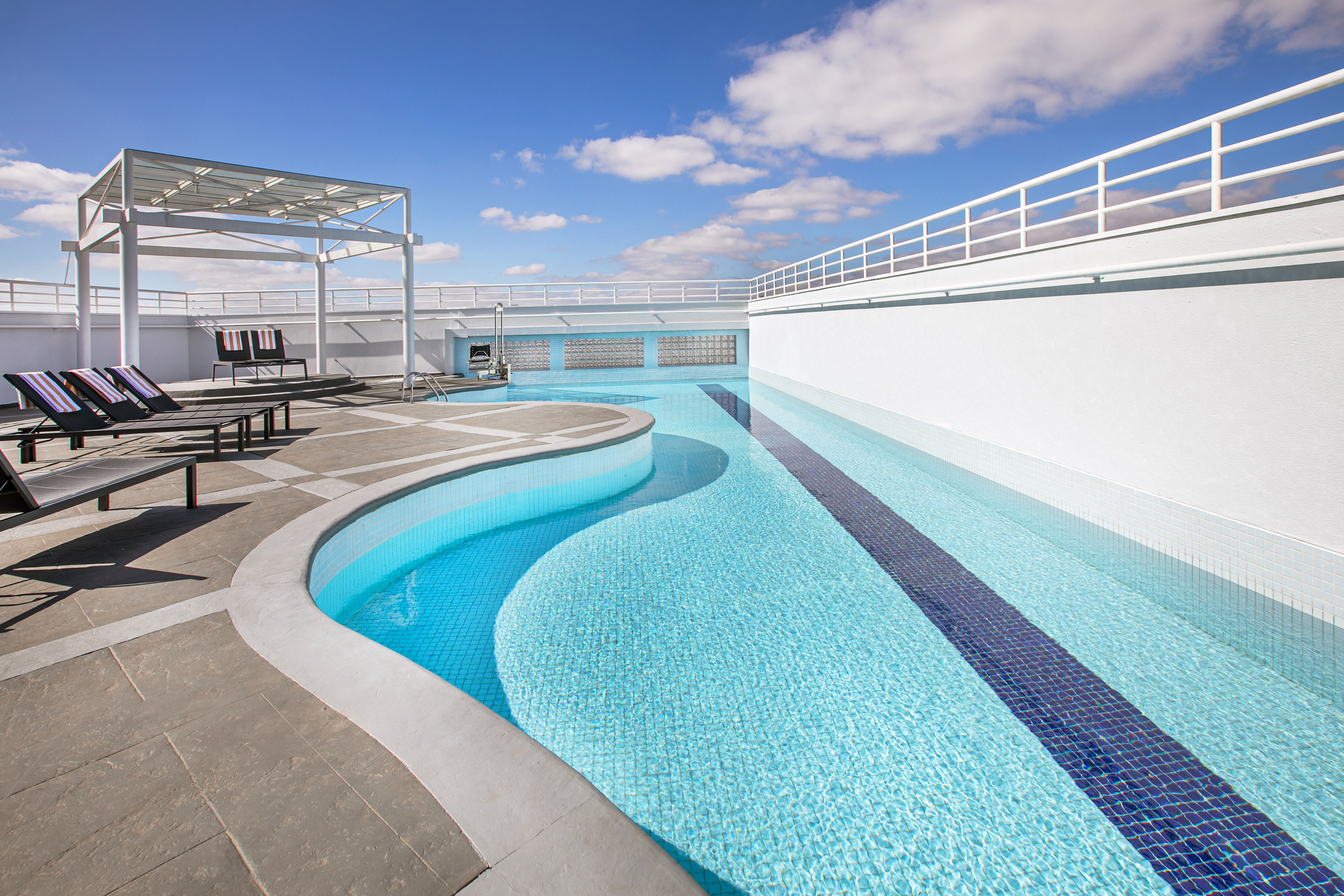 Rooftop pool with blue tiled water, sun loungers, and white pergola under sunny sky.