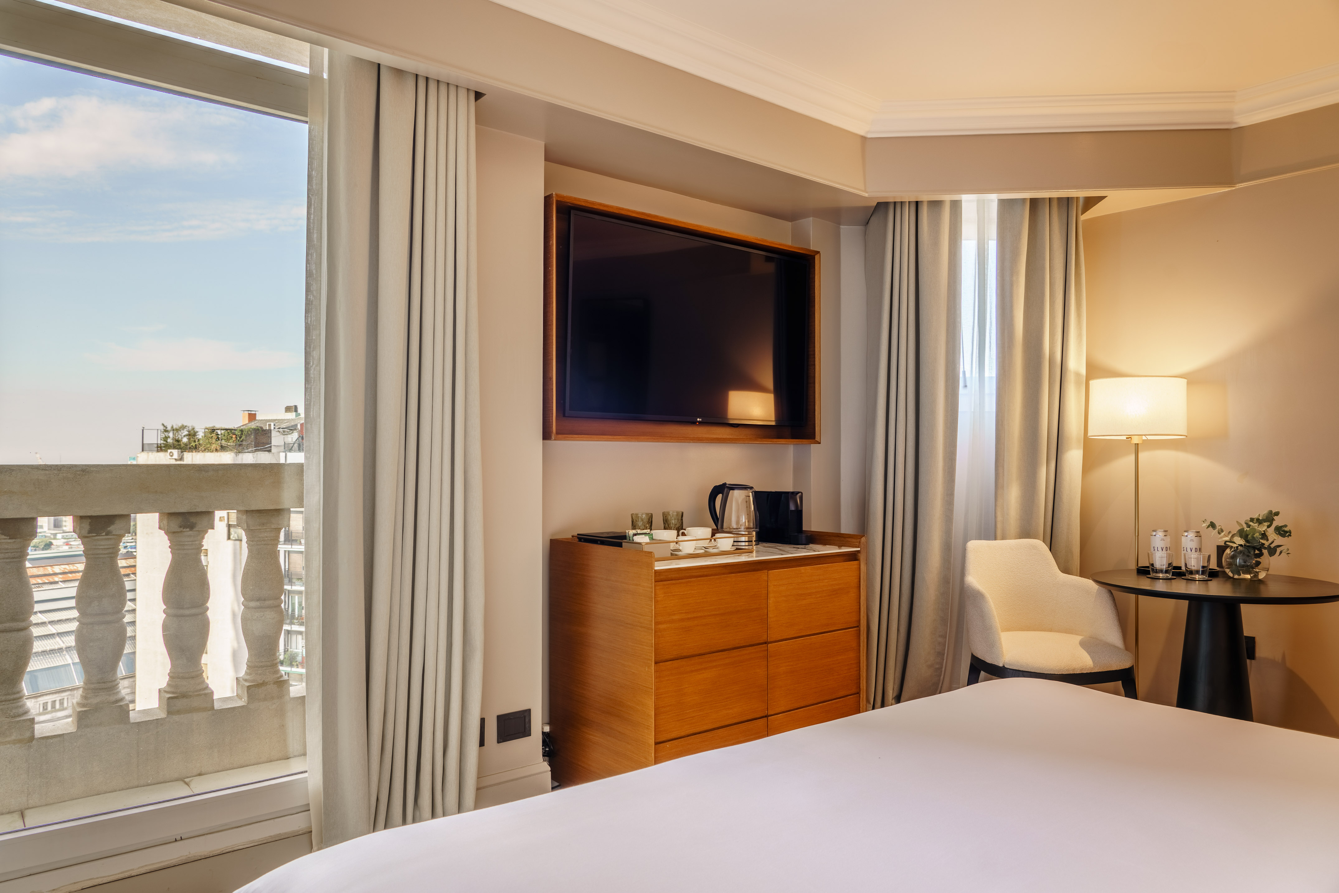 Modern hotel room with balcony city view, TV, bed, and tea station.