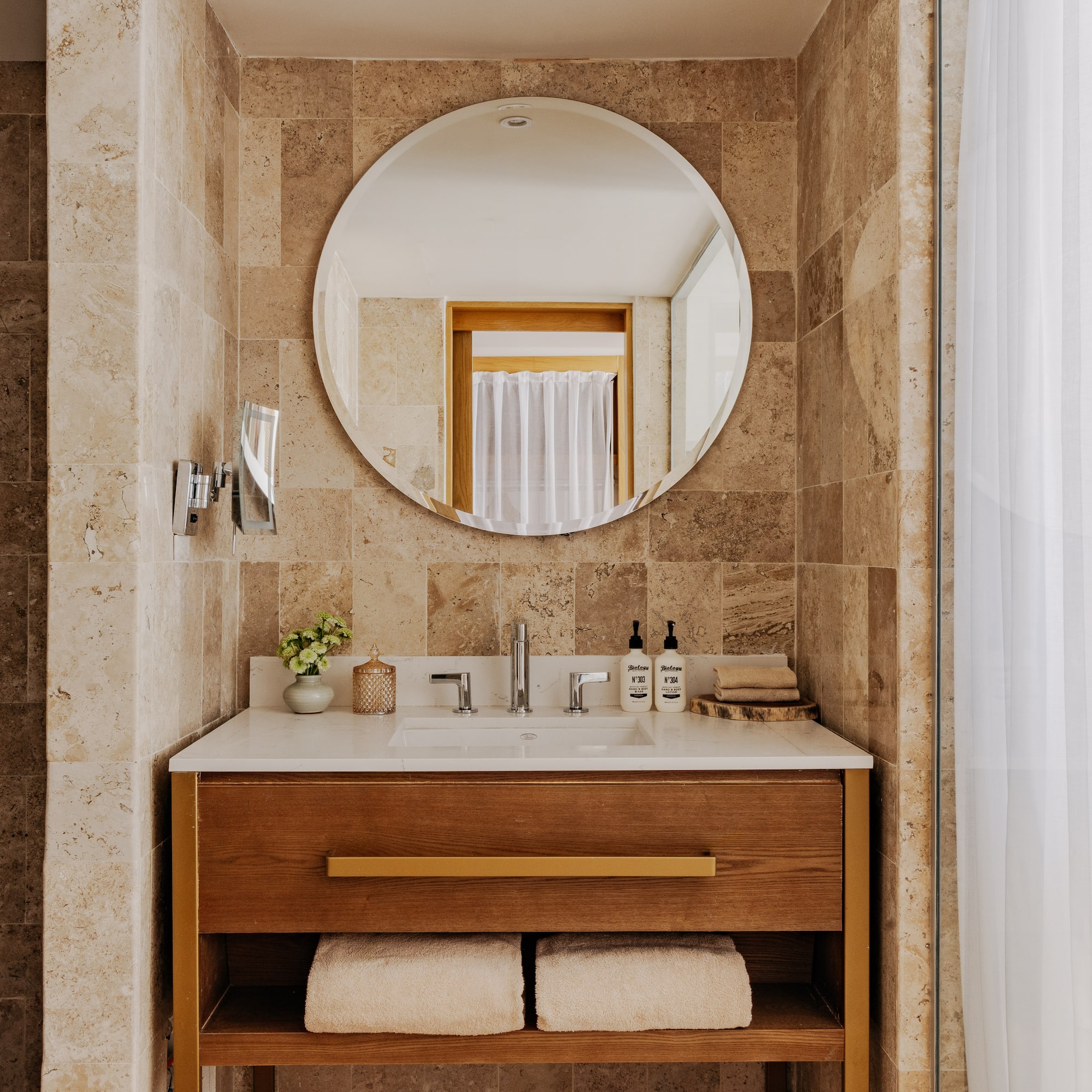 Modern bathroom vanity with wood cabinet, round mirror, stone tiles, white sink.
