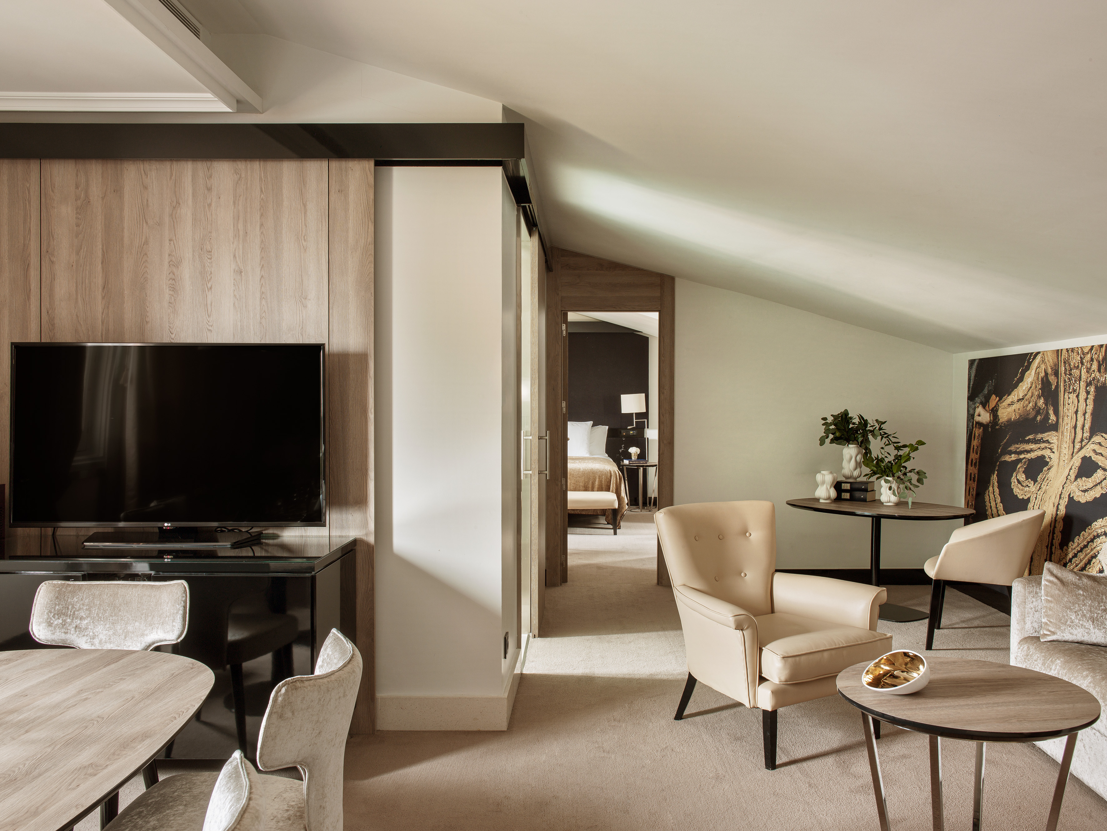 Modern hotel suite living room with TV, elegant beige furniture, and bedroom.