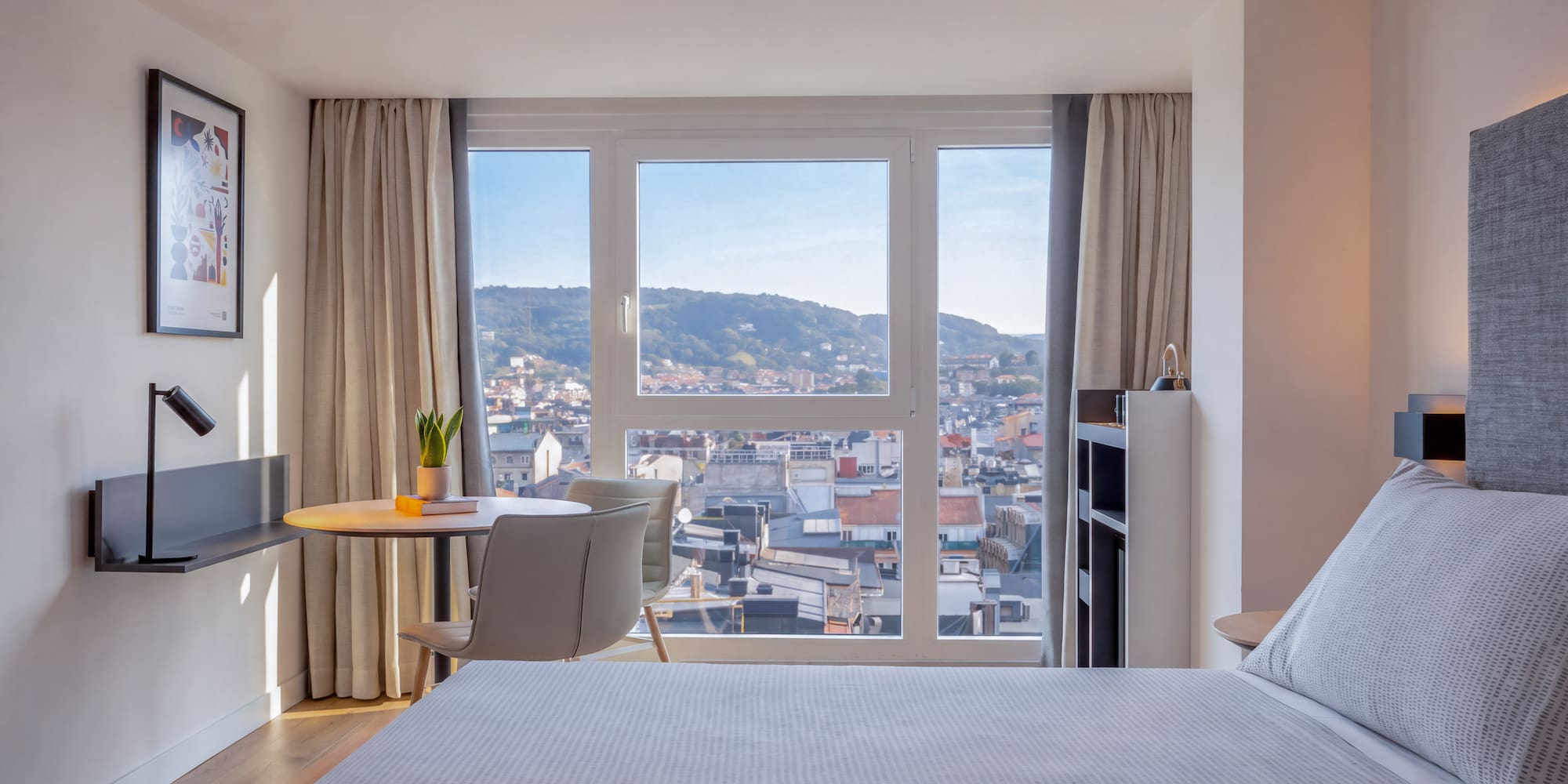 Dormitorio de hotel moderno con cama, escritorio y vista panorámica a la ciudad.