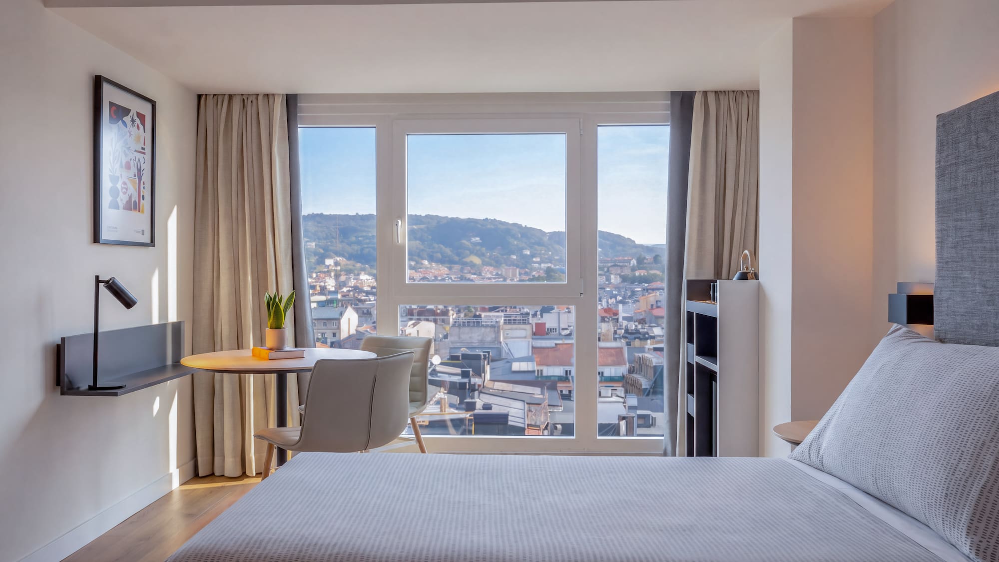 Dormitorio de hotel moderno con cama, escritorio y vista panorámica a la ciudad.