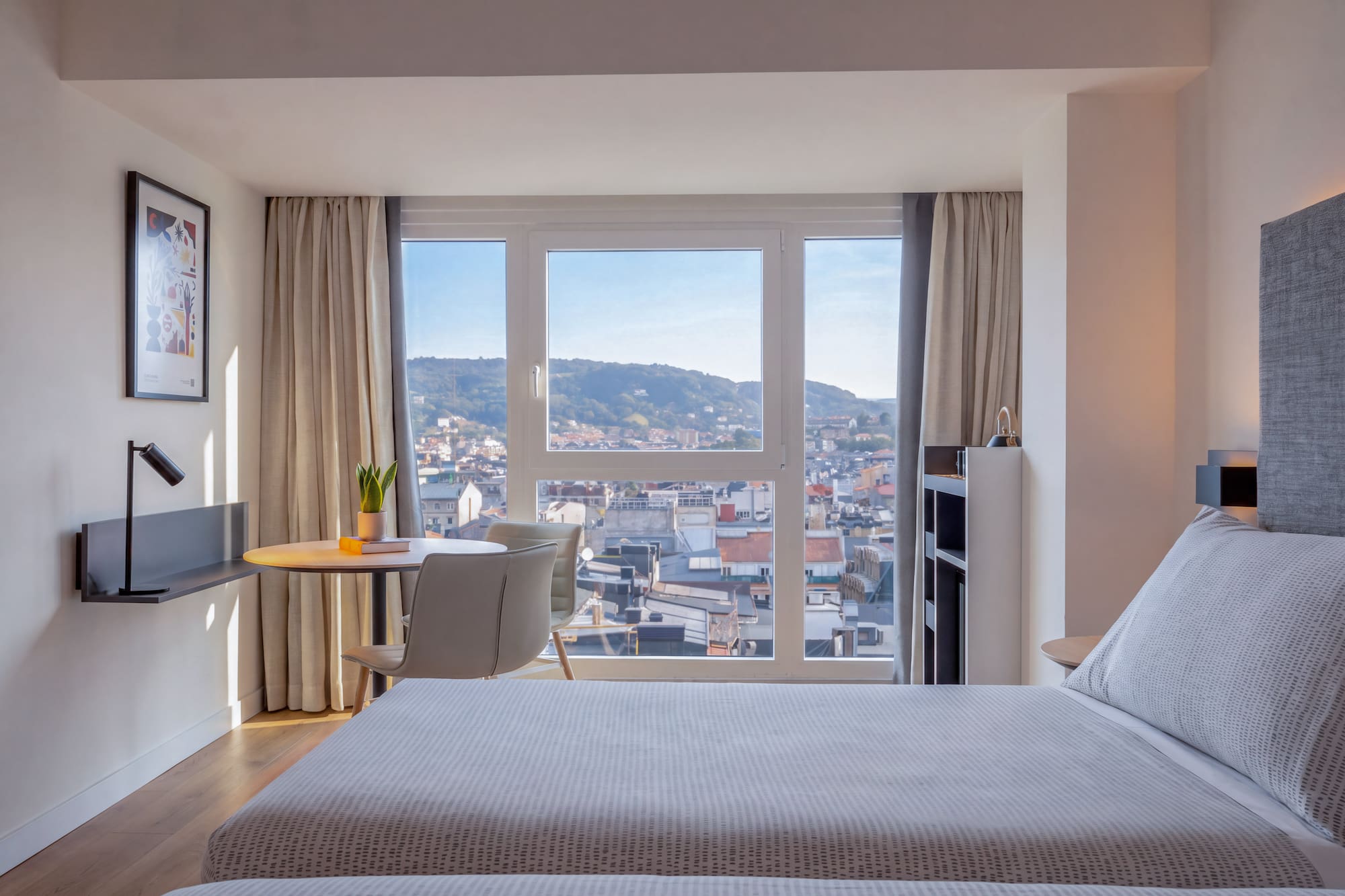 Dormitorio de hotel moderno con cama, escritorio y vista panorámica a la ciudad.