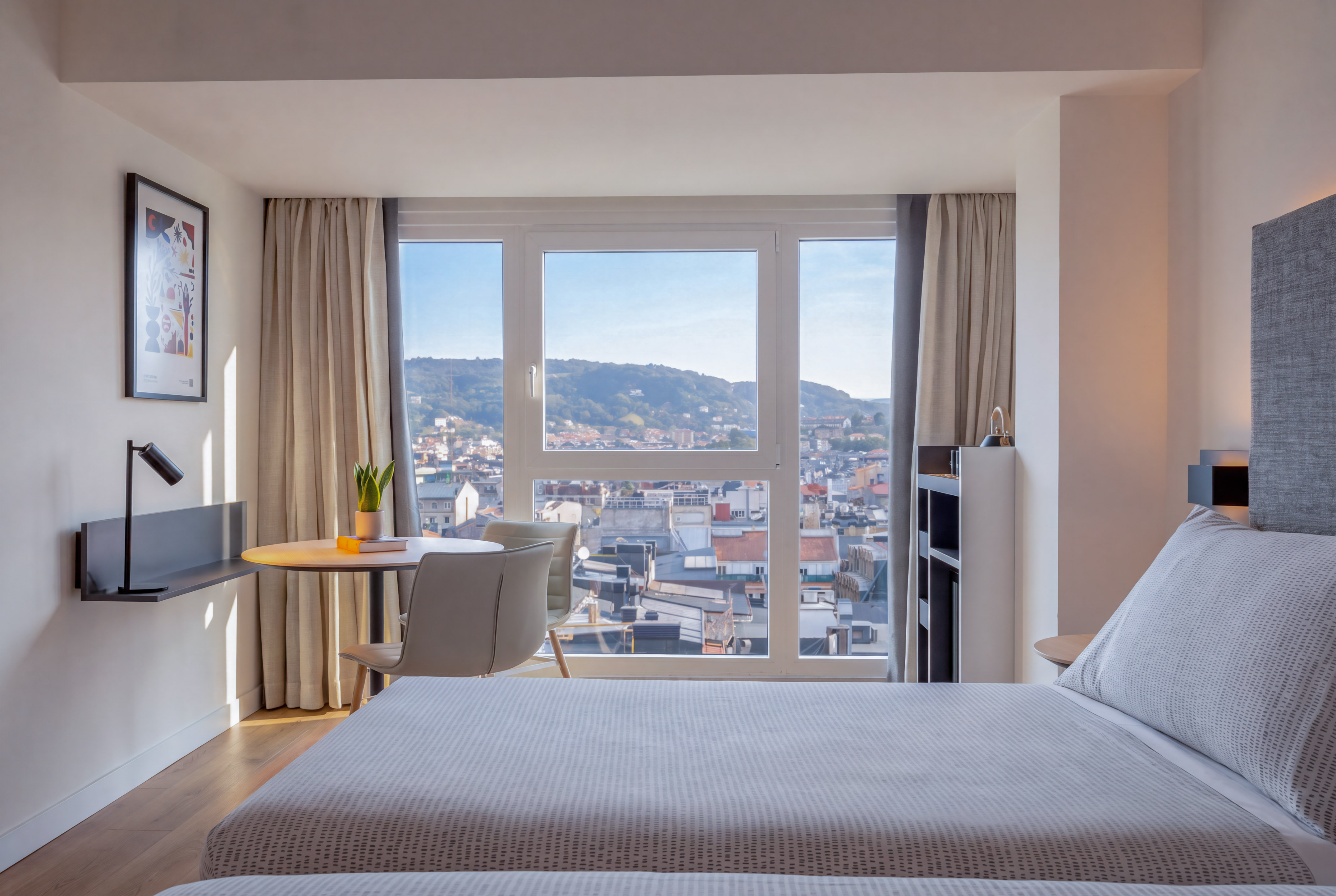 Dormitorio de hotel moderno con cama, escritorio y vista panorámica a la ciudad.