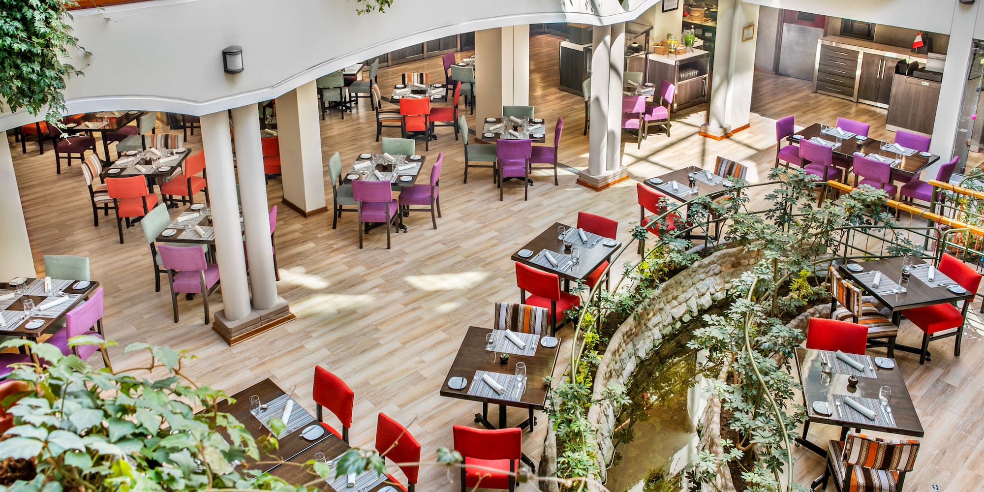 Colorful restaurant atrium with dining tables, chairs, plants, and pond.