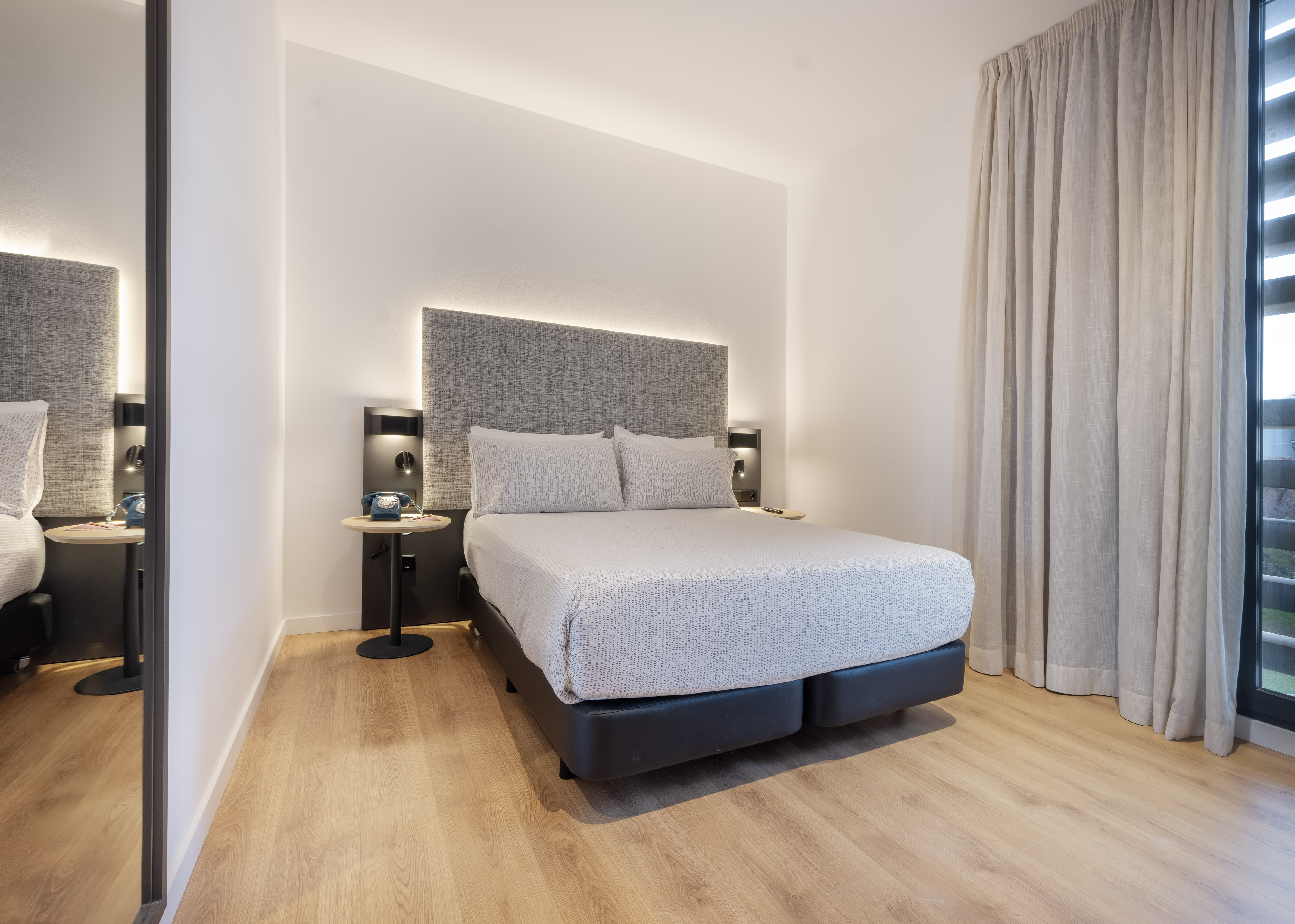 Modern hotel room with bed, backlit grey headboard, wood floor, and window.