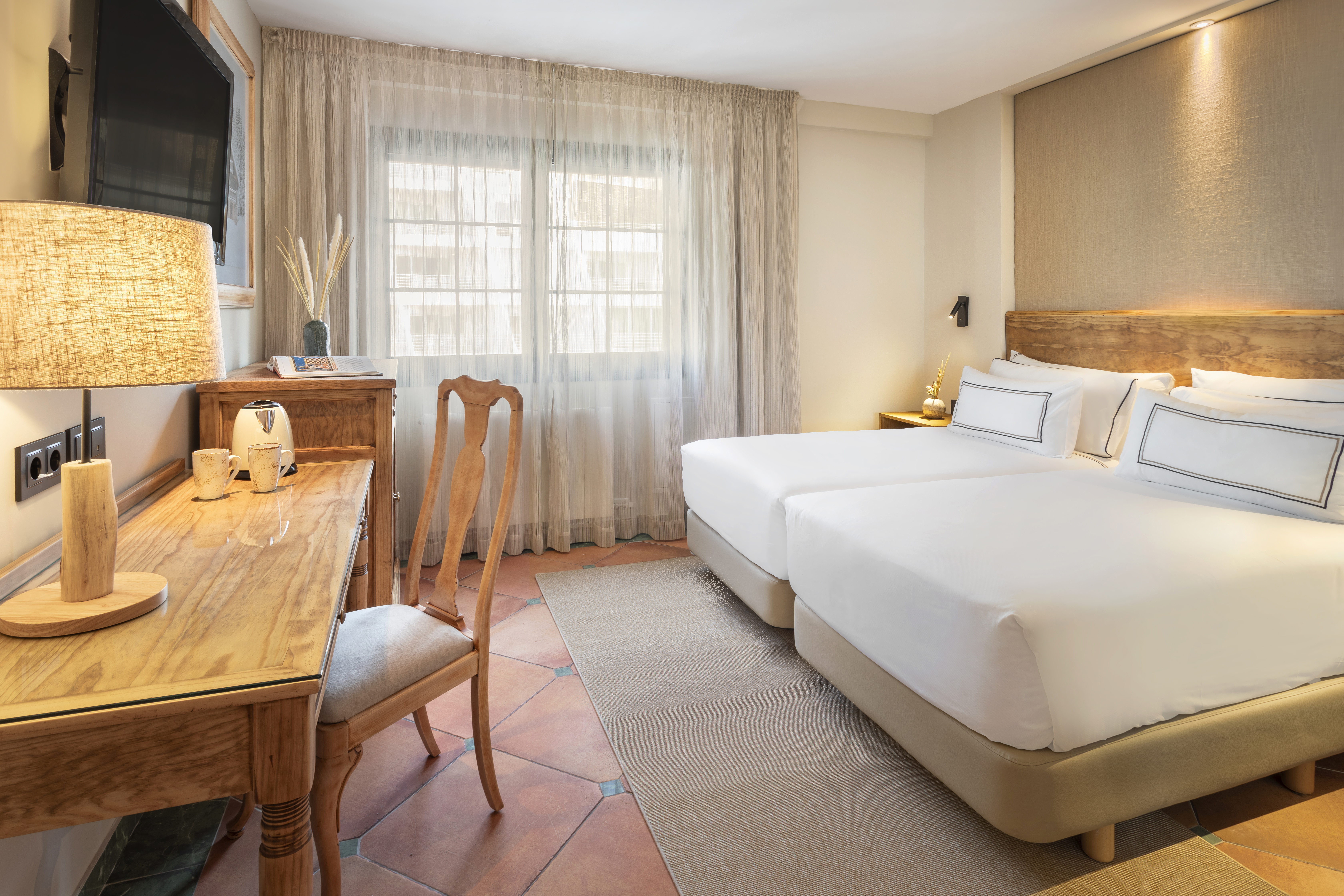 Bright hotel bedroom with twin beds, wooden desk, TV, and large window.