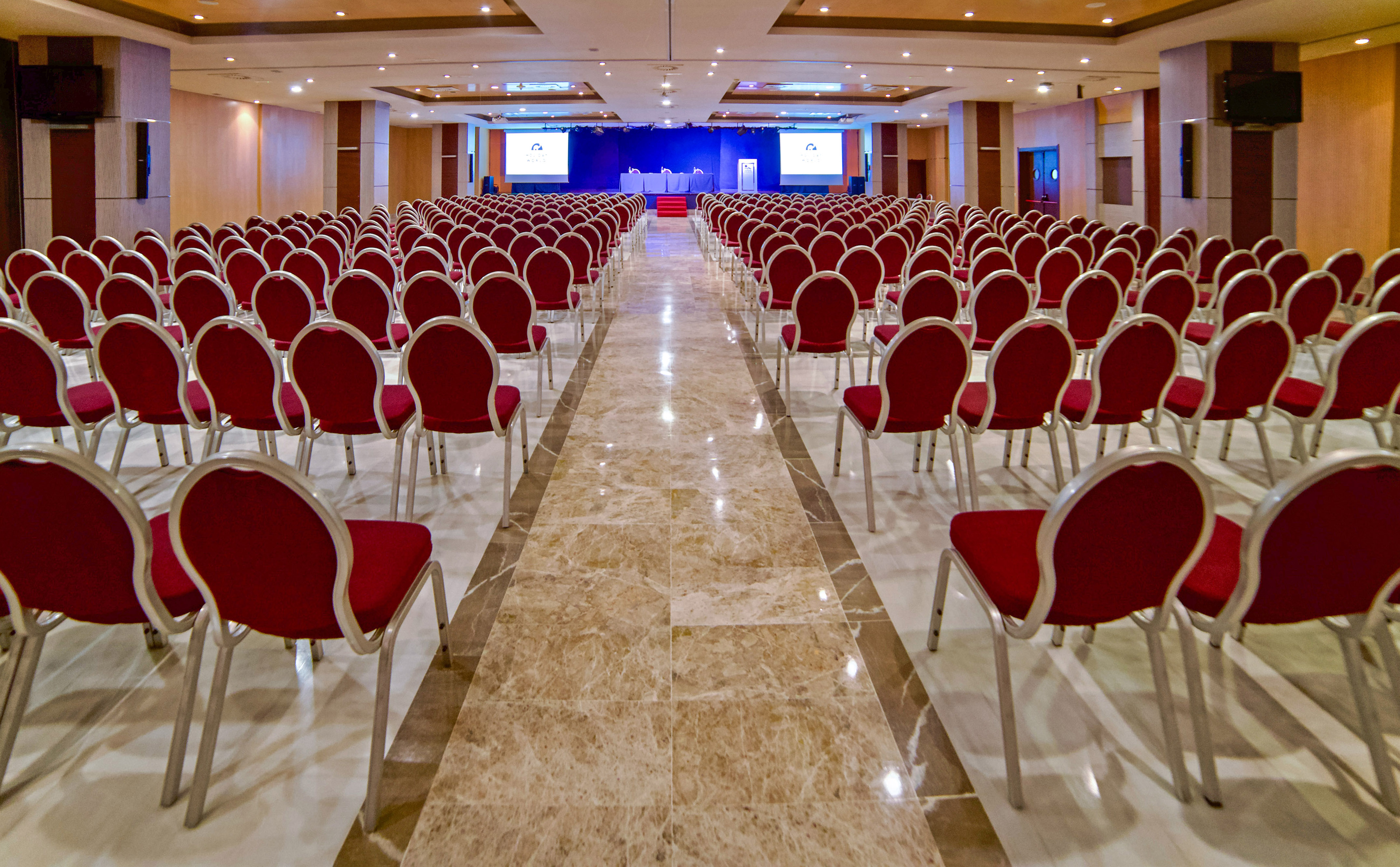 Spacious conference room with rows of red chairs facing a stage with screens.