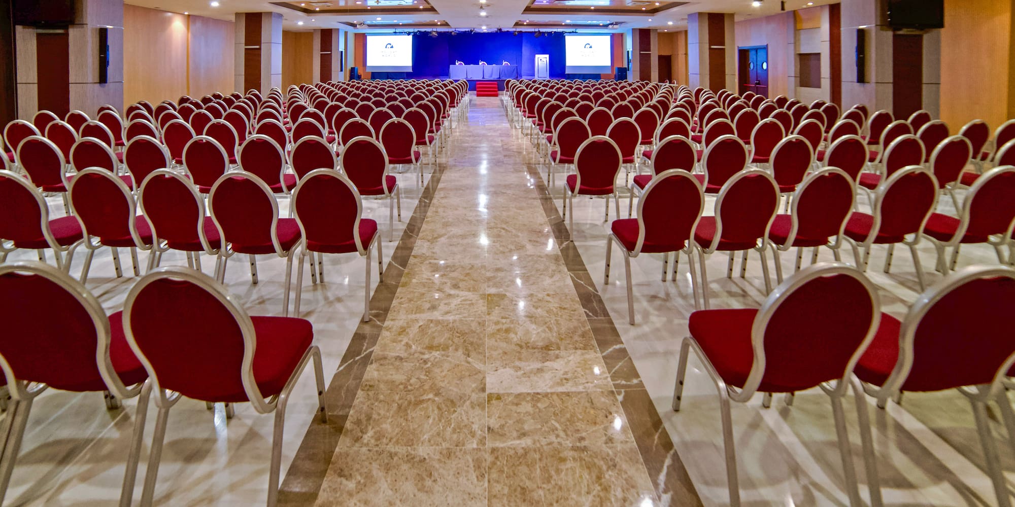 Spacious conference room with rows of red chairs facing a stage with screens.