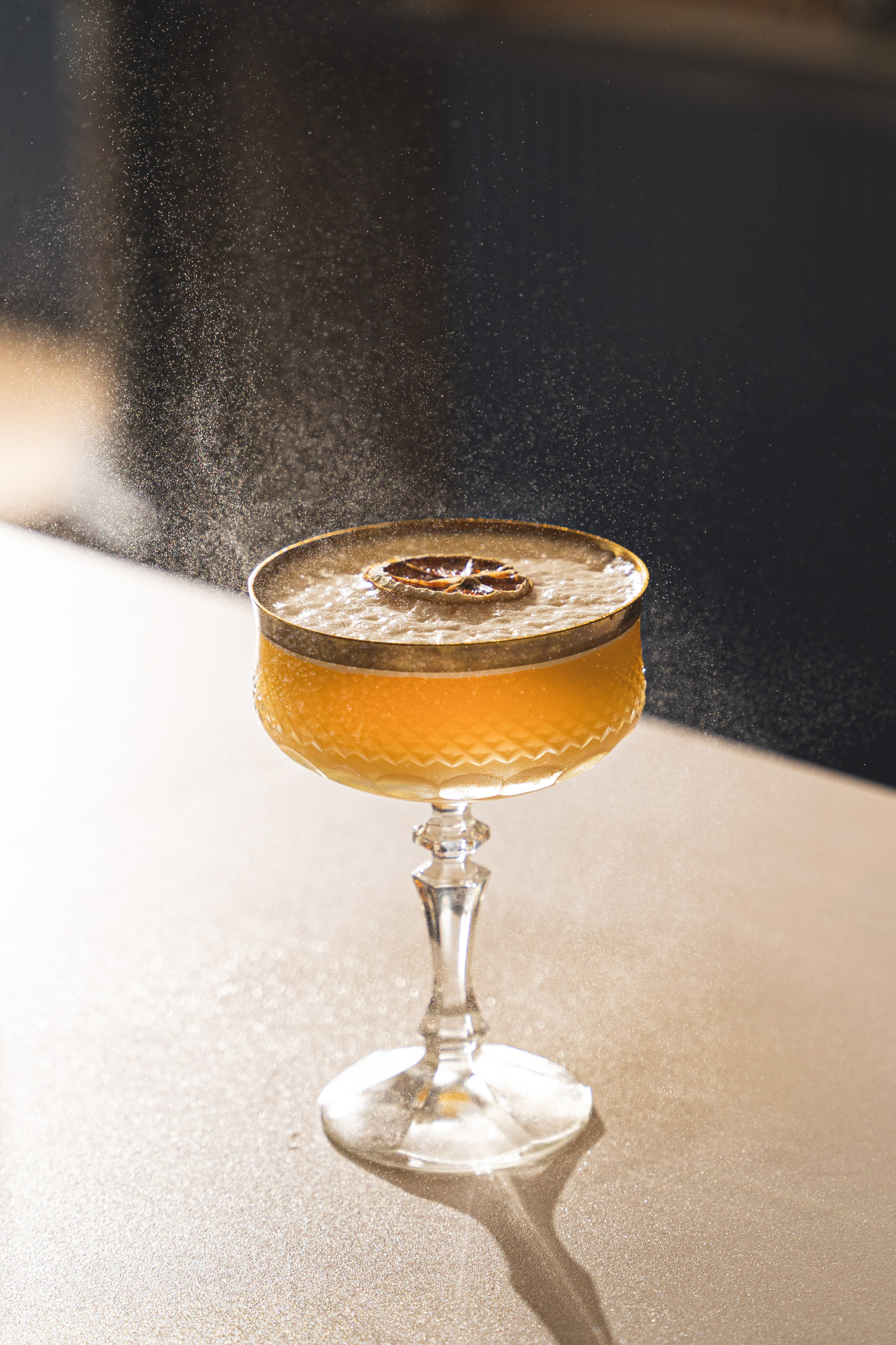 Orange cocktail in coupe glass with dried citrus garnish and fine spray.