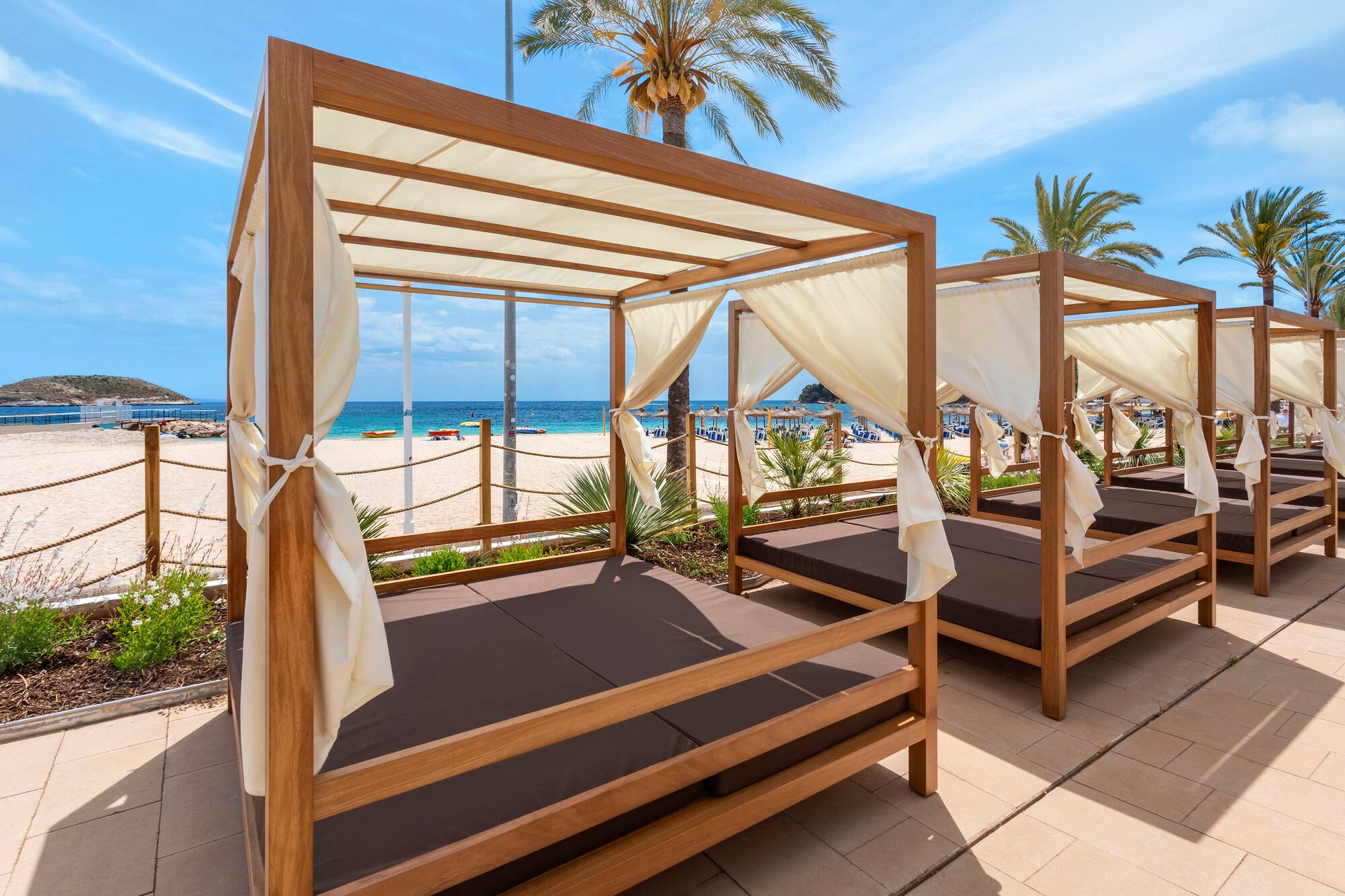 Resort beach cabanas with cream curtains, brown cushions, ocean, and palm trees.