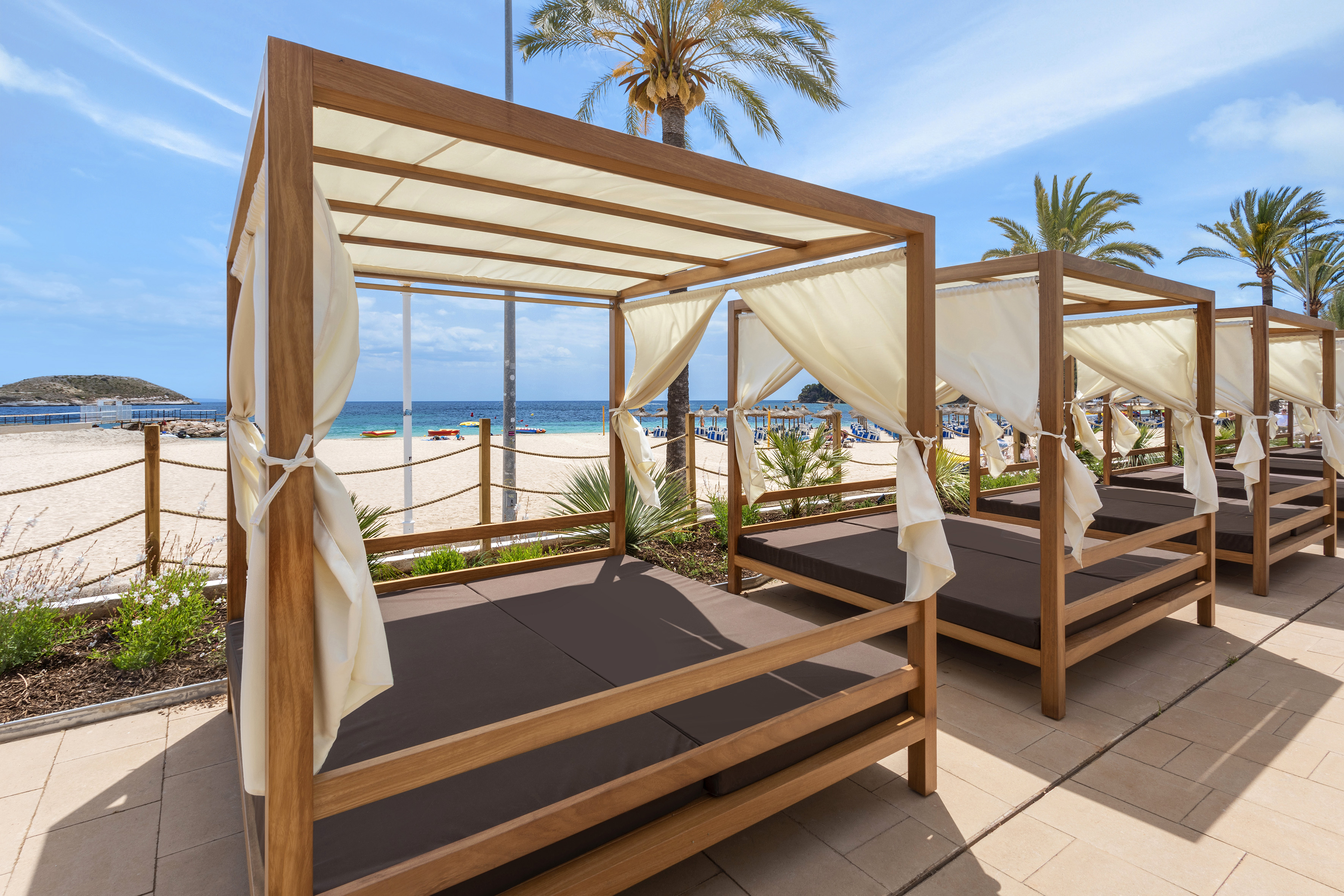 Resort beach cabanas with cream curtains, brown cushions, ocean, and palm trees.