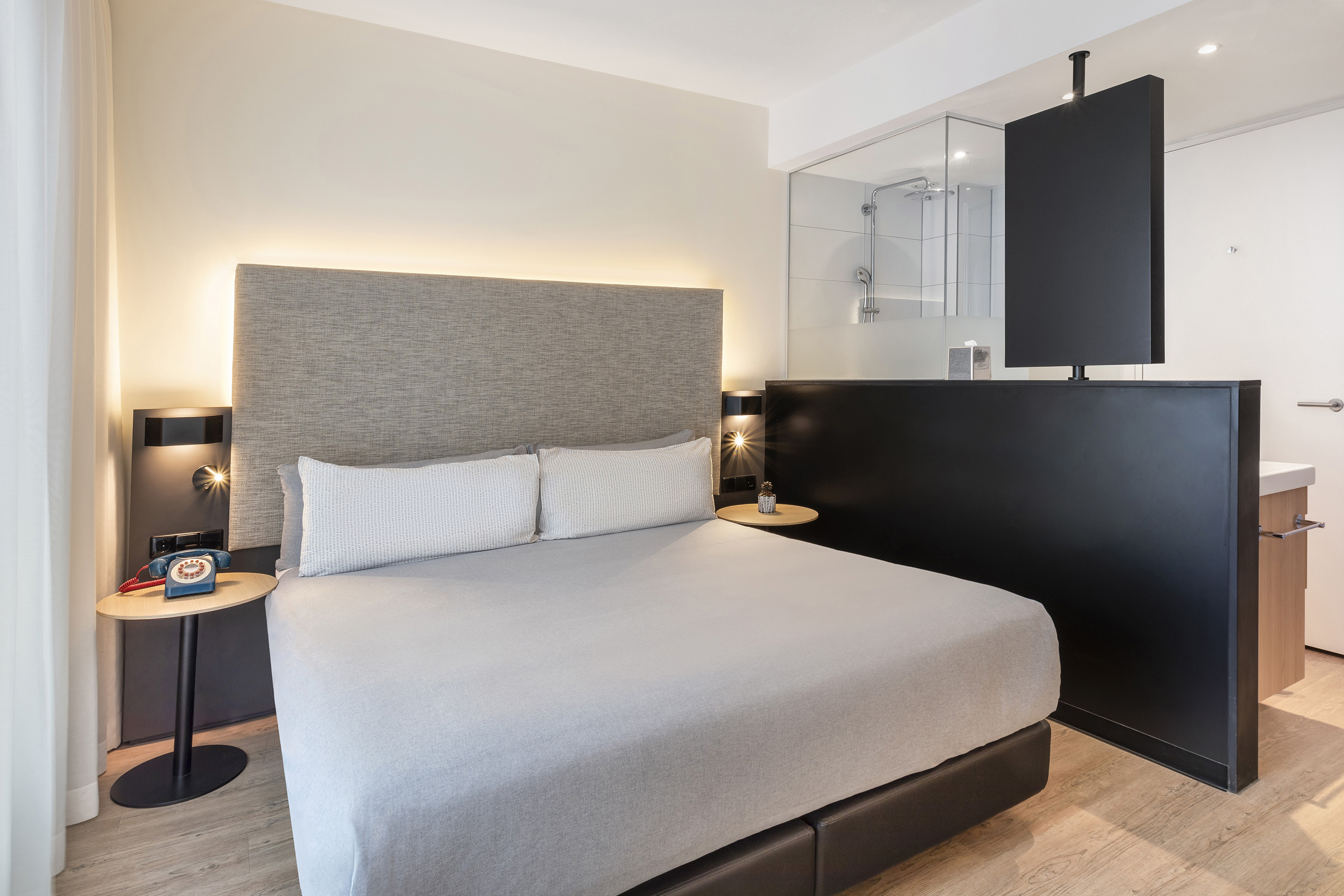 Modern hotel room with king bed, backlit headboard, glass shower, and partition TV.