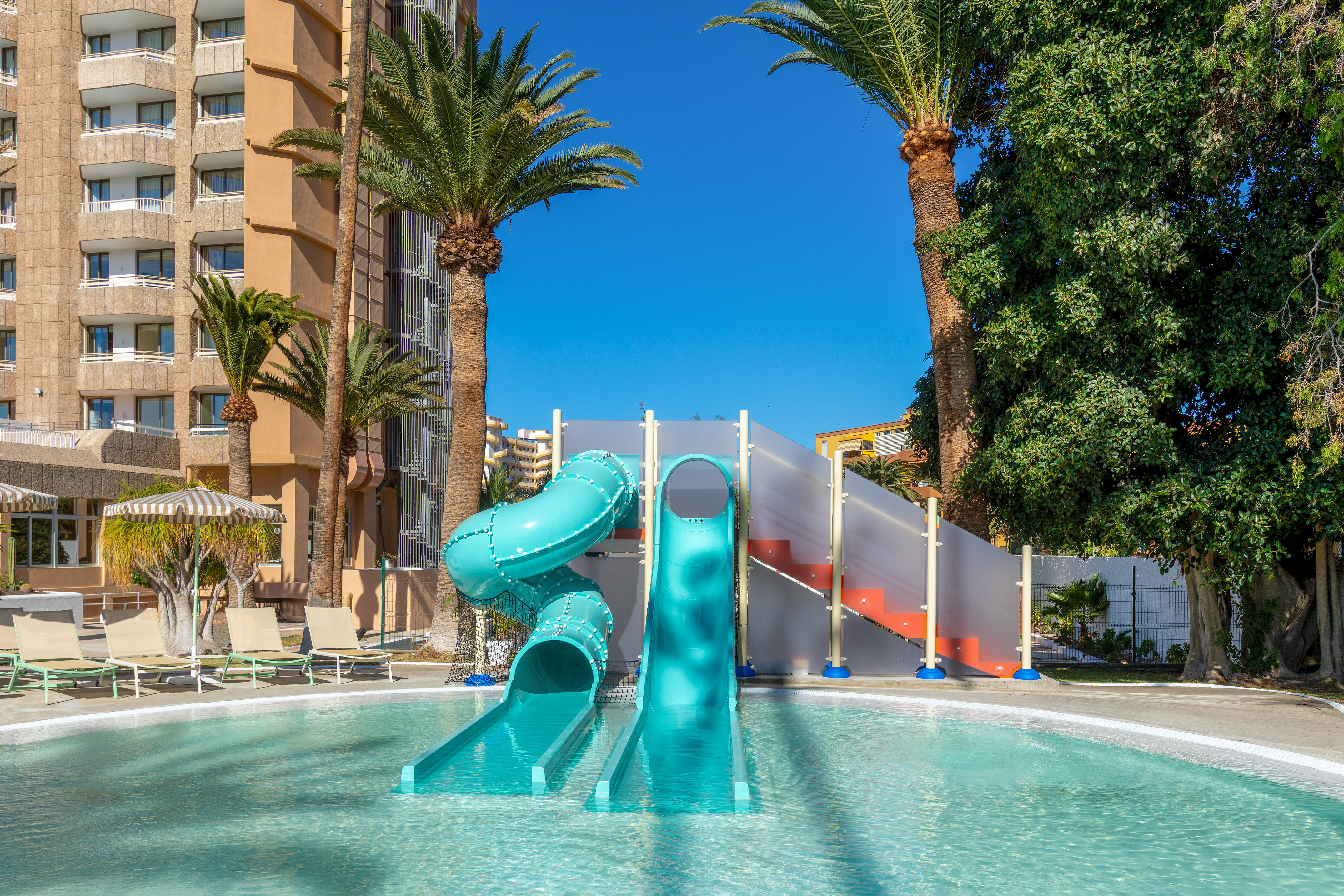 Hotel pool with two blue water slides, palm trees, and resort building.