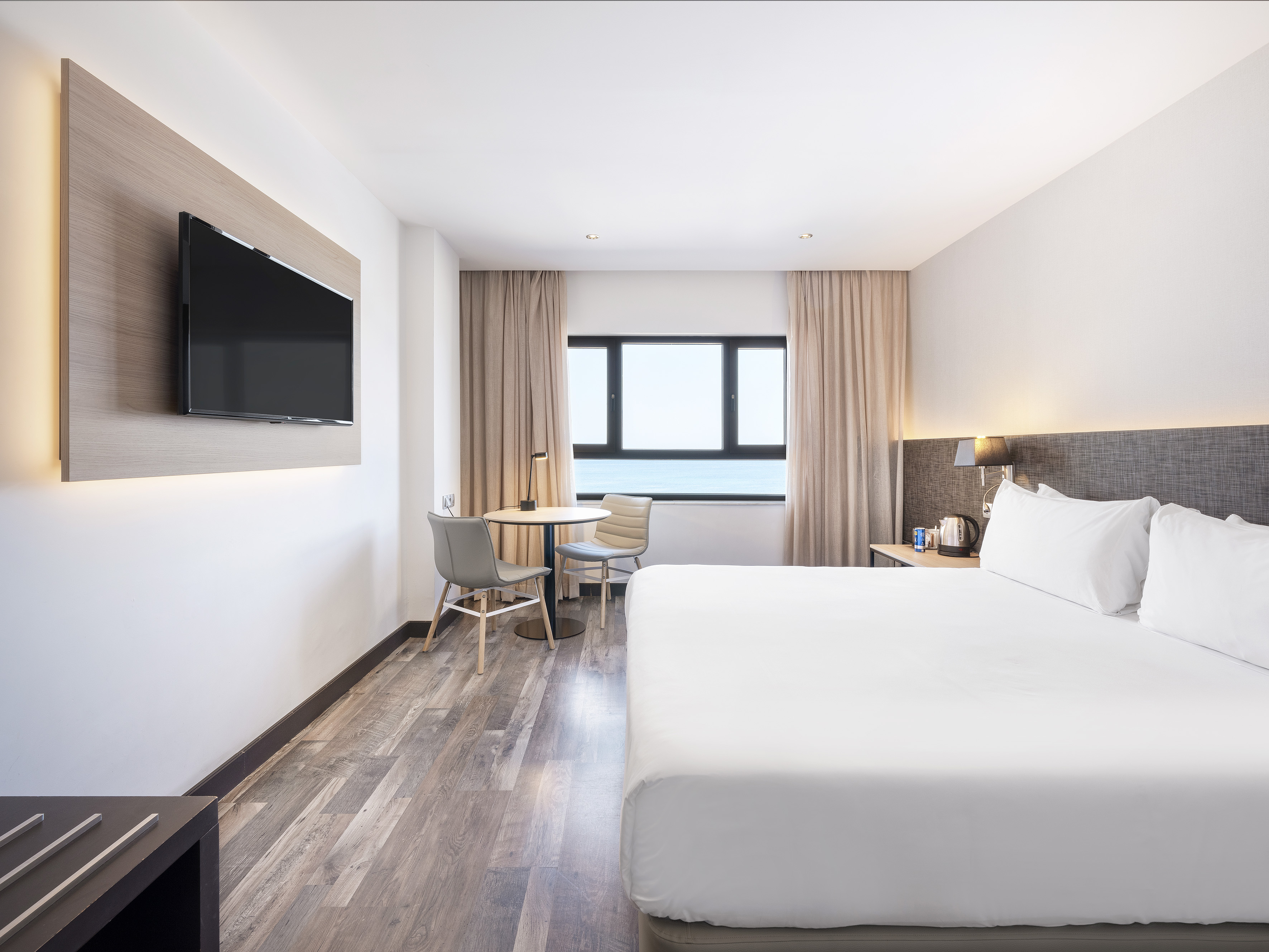 Modern hotel room with king bed, ocean view, wall TV, and small table.