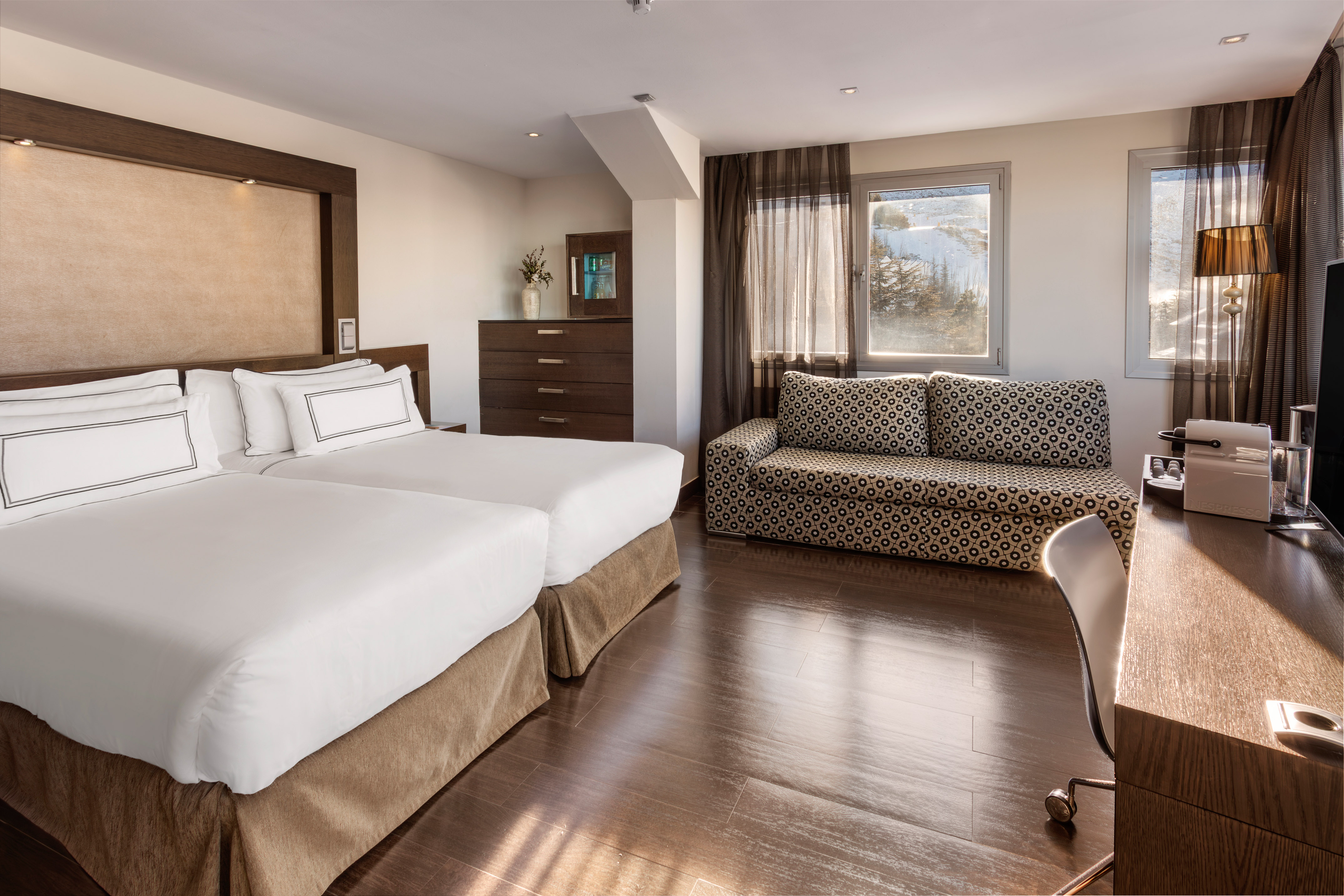 Cozy hotel room with twin beds, patterned sofa, desk, and snowy mountain view.