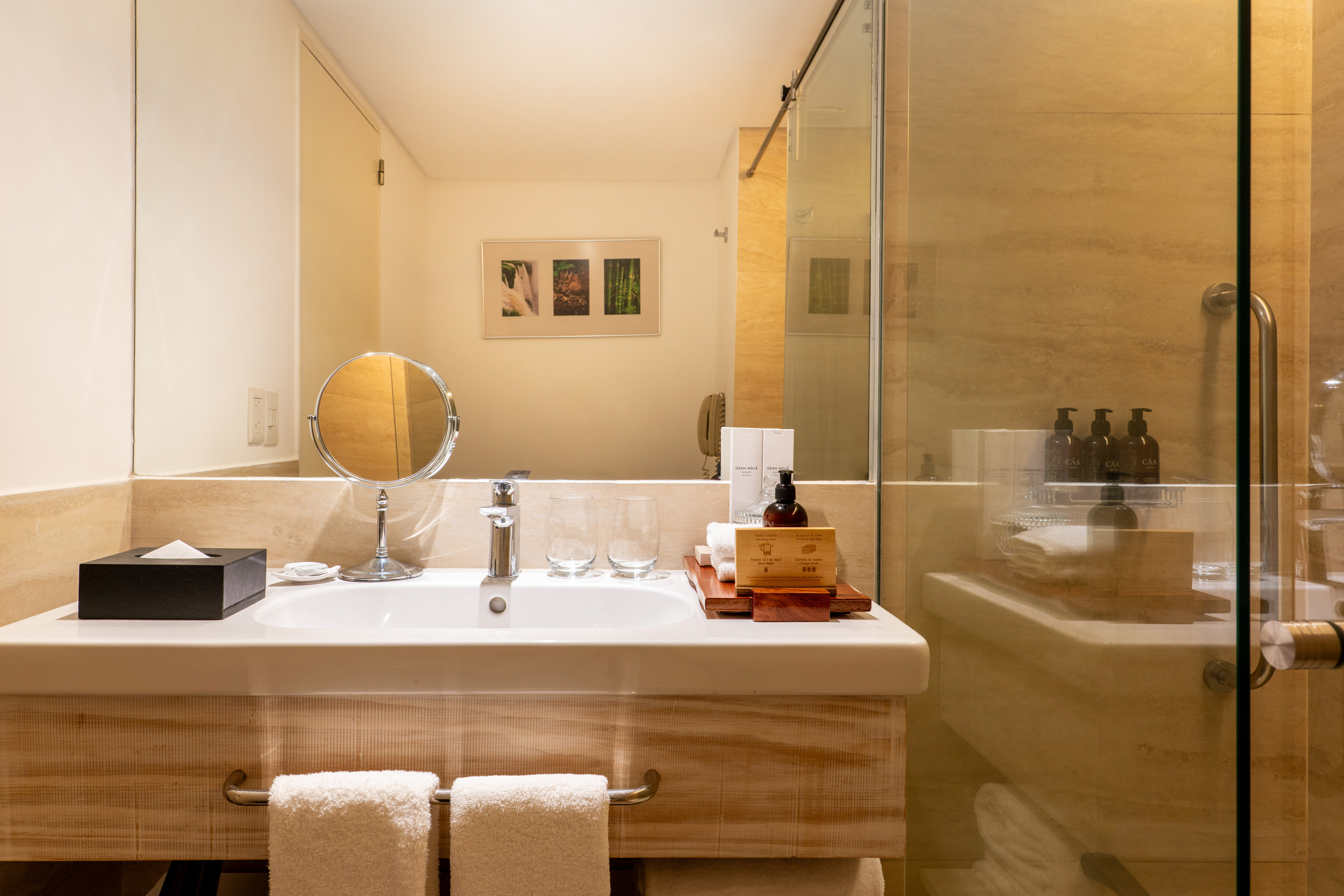 Beige hotel bathroom vanity with white sink, mirror, glass shower, and towels.