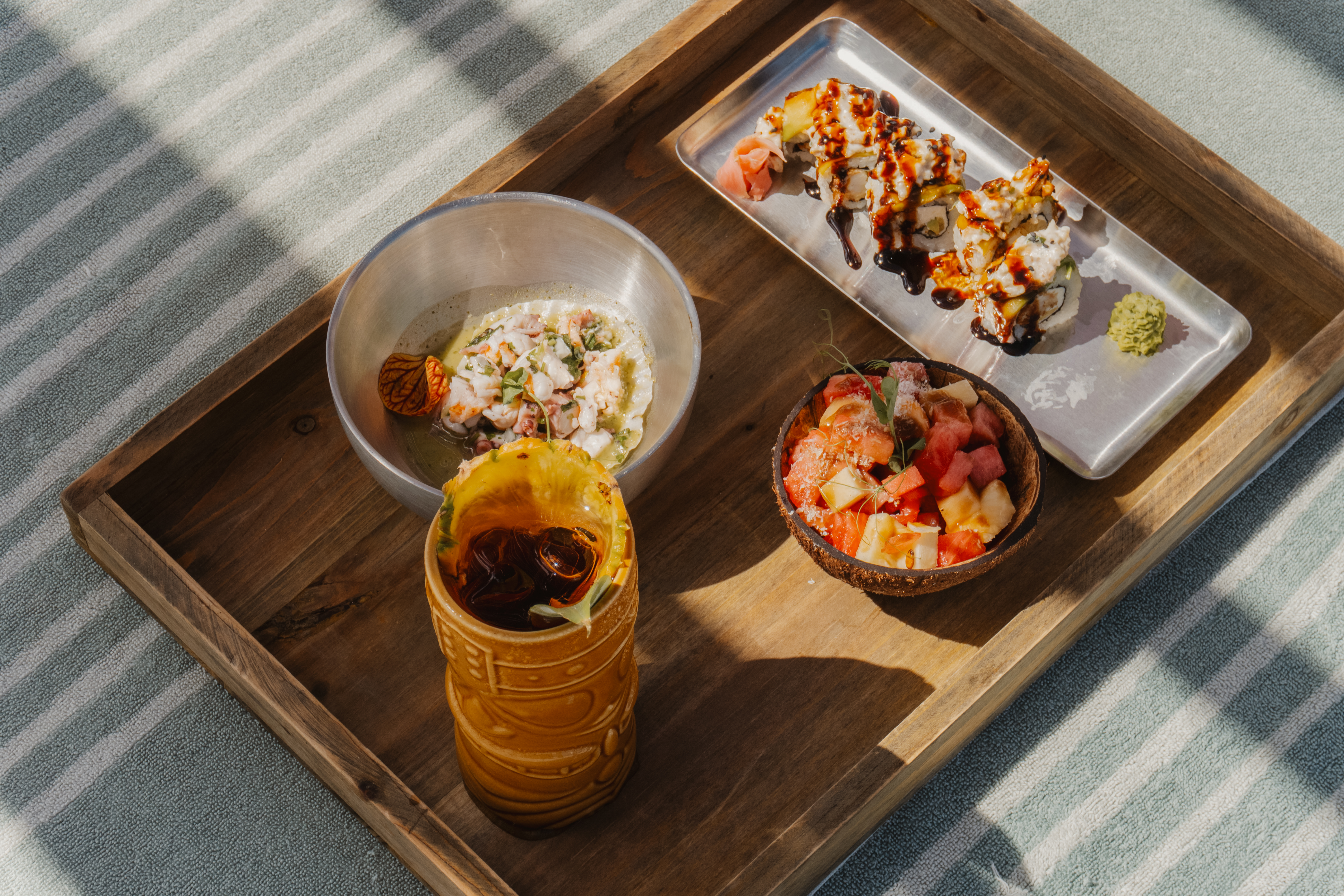 Tropical meal on wooden tray: sushi, ceviche, fruit salad, tiki drink.