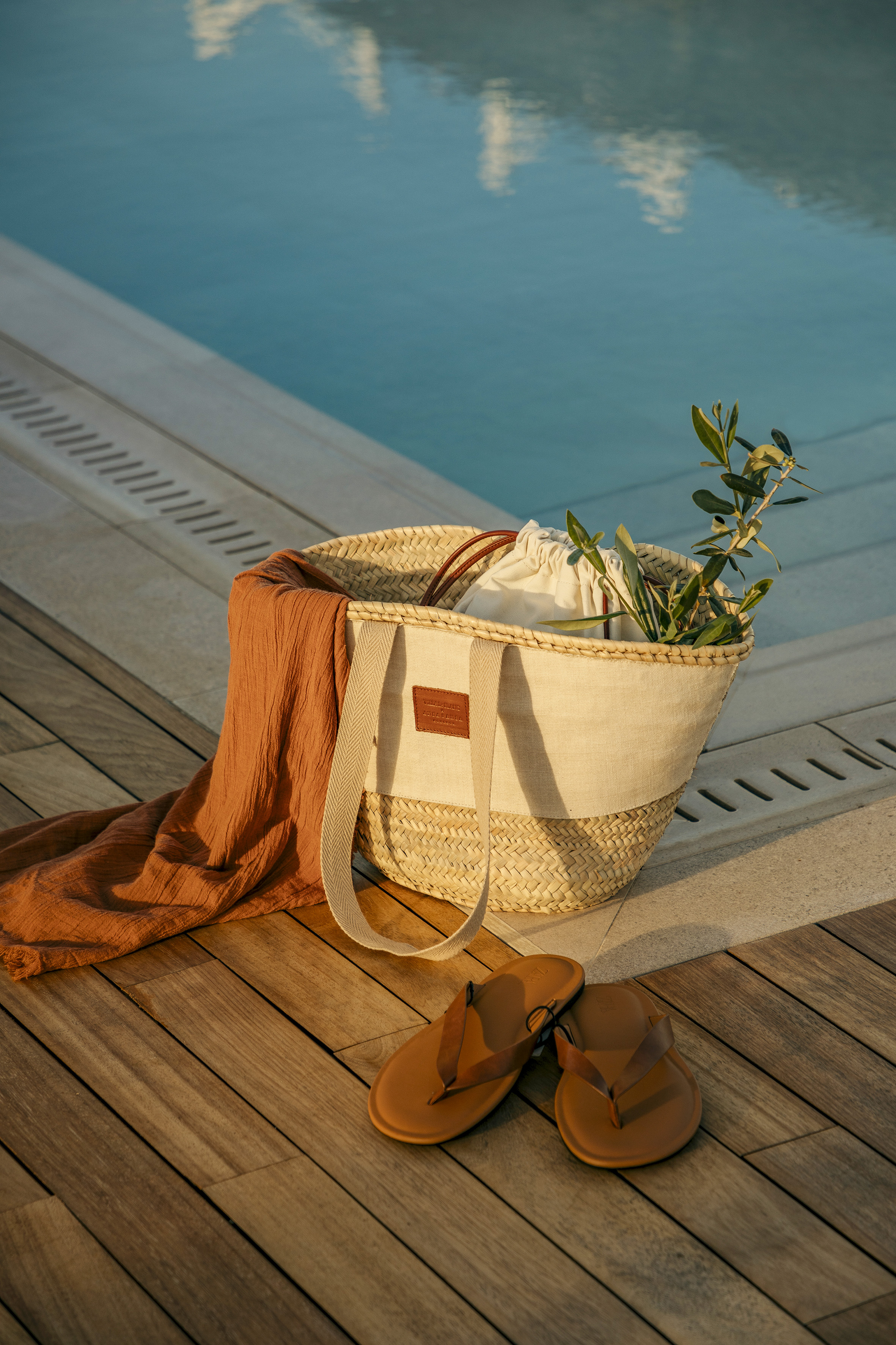 Poolside straw bag, orange cover-up, brown flip-flops on wooden deck.