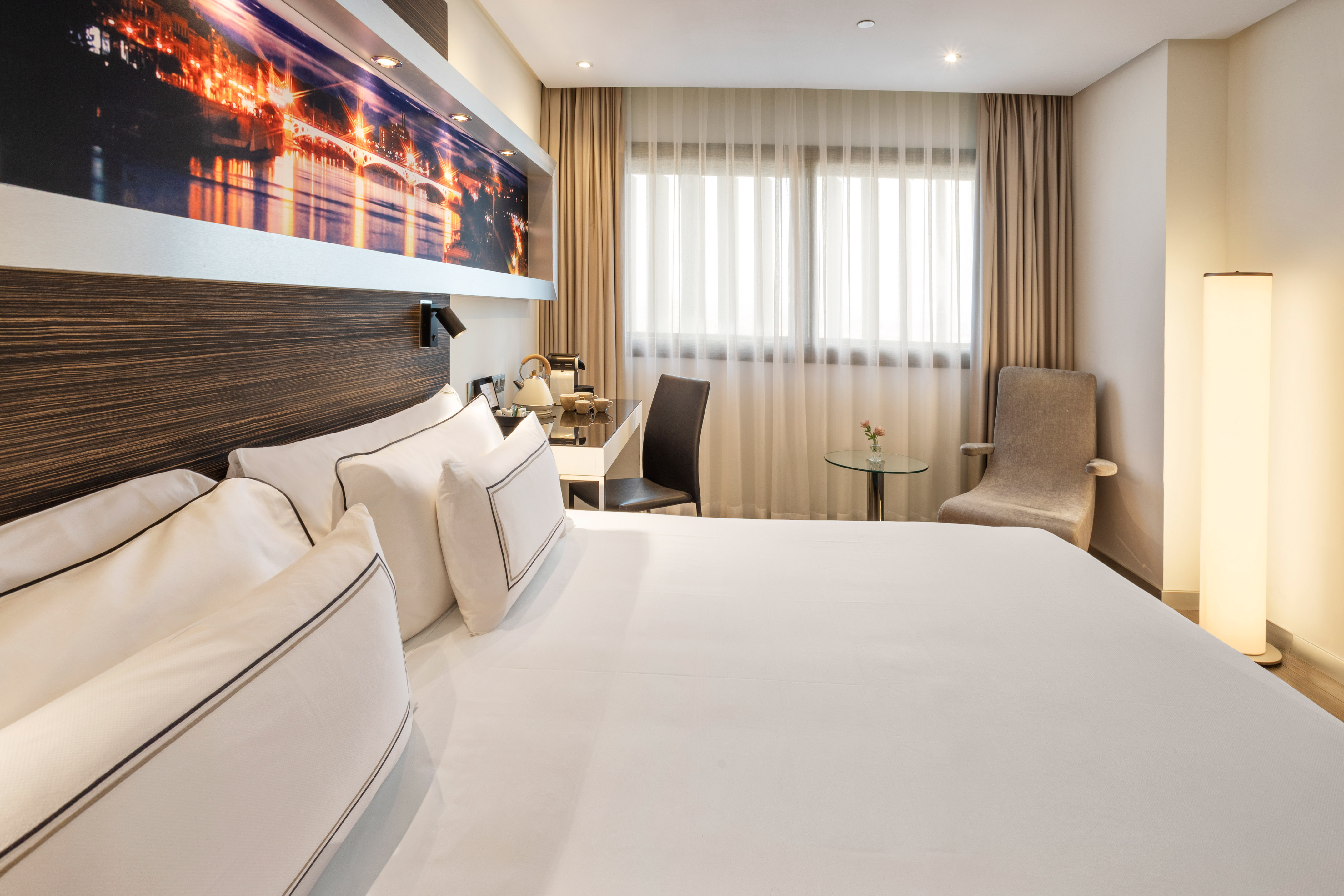 Modern hotel room with king bed, desk area, armchair, and cityscape artwork.