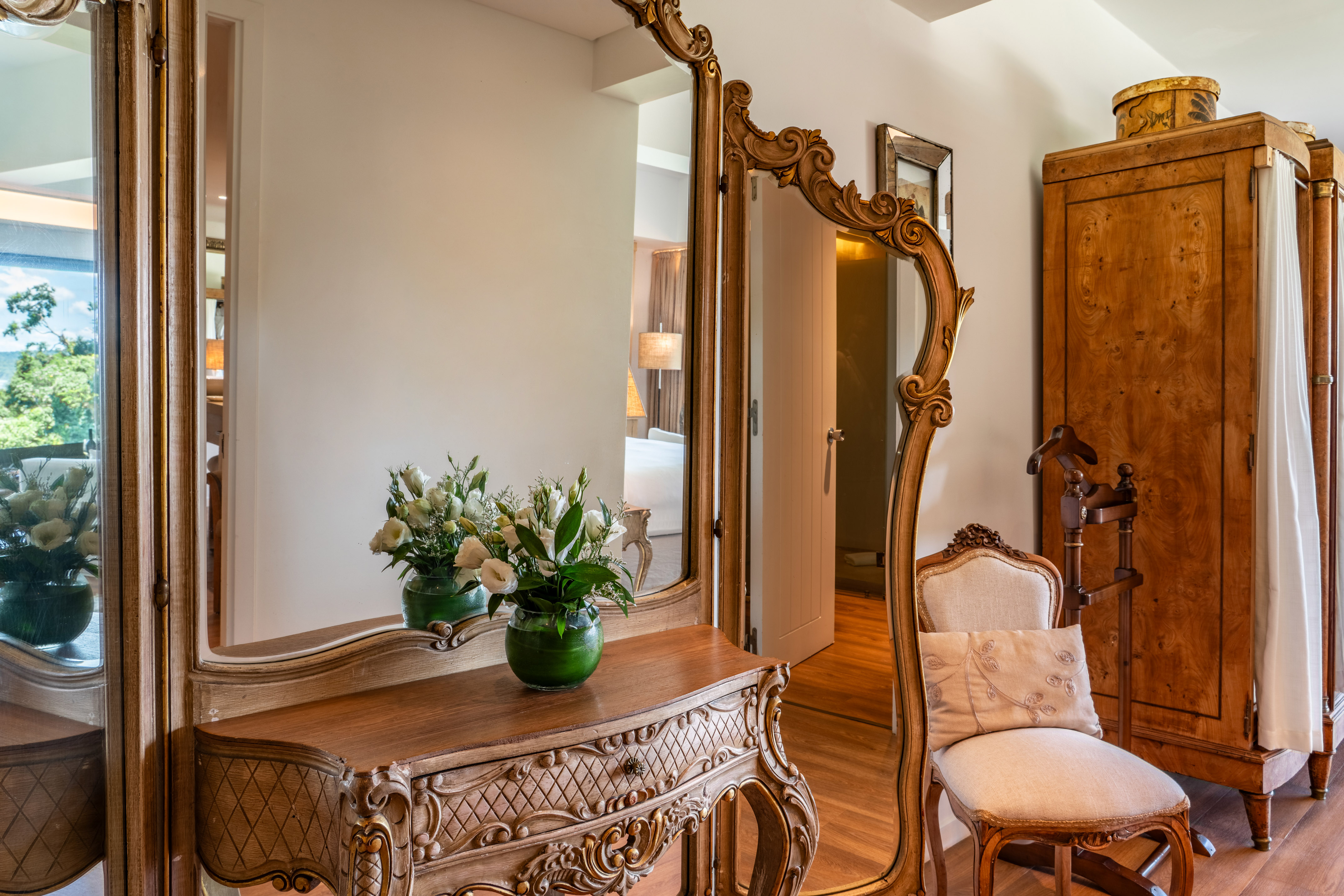 Ornate gold mirror, carved vanity, flowers, and antique wardrobe in bedroom.
