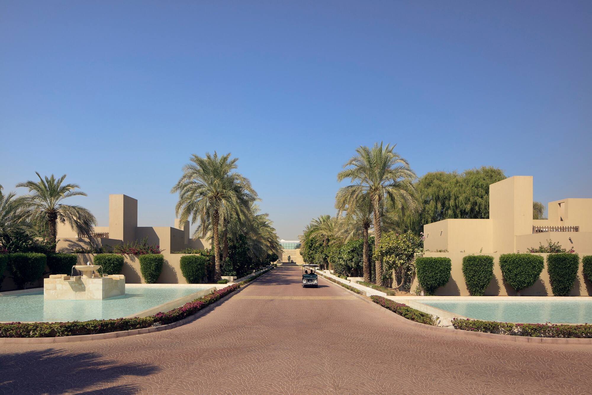 Resort entrance with palm trees, modern sand-colored buildings, and pools.