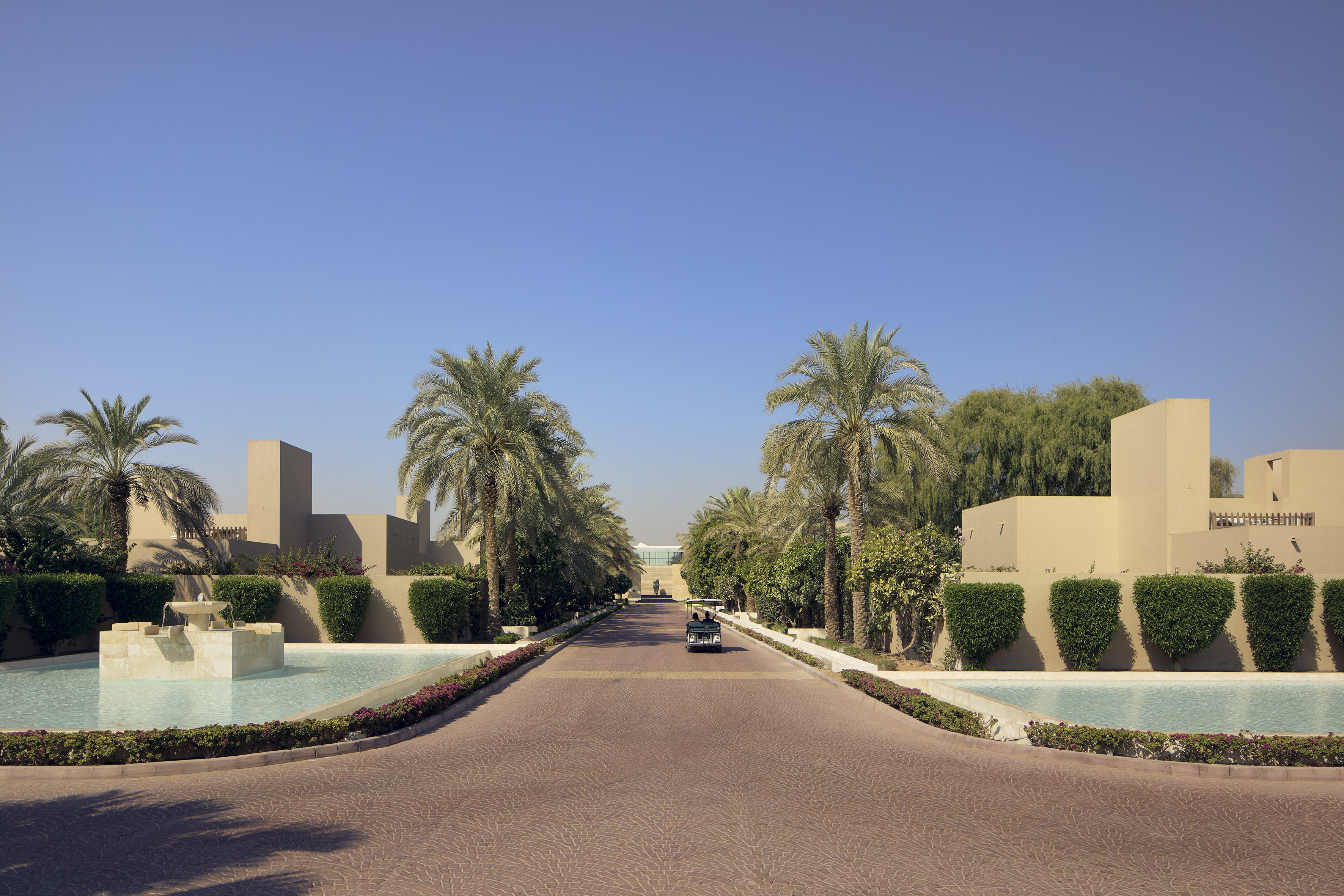 Resort entrance with palm trees, modern sand-colored buildings, and pools.