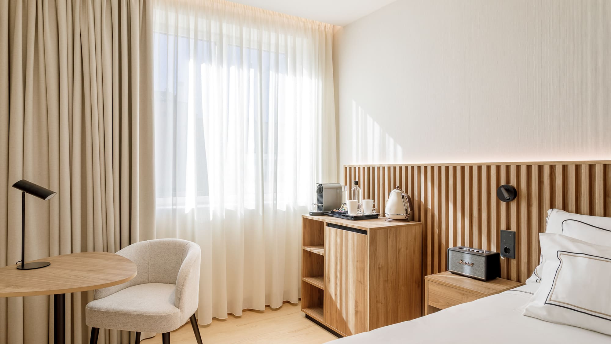 Modern hotel room with wooden headboard, minibar, coffee machine, and table.