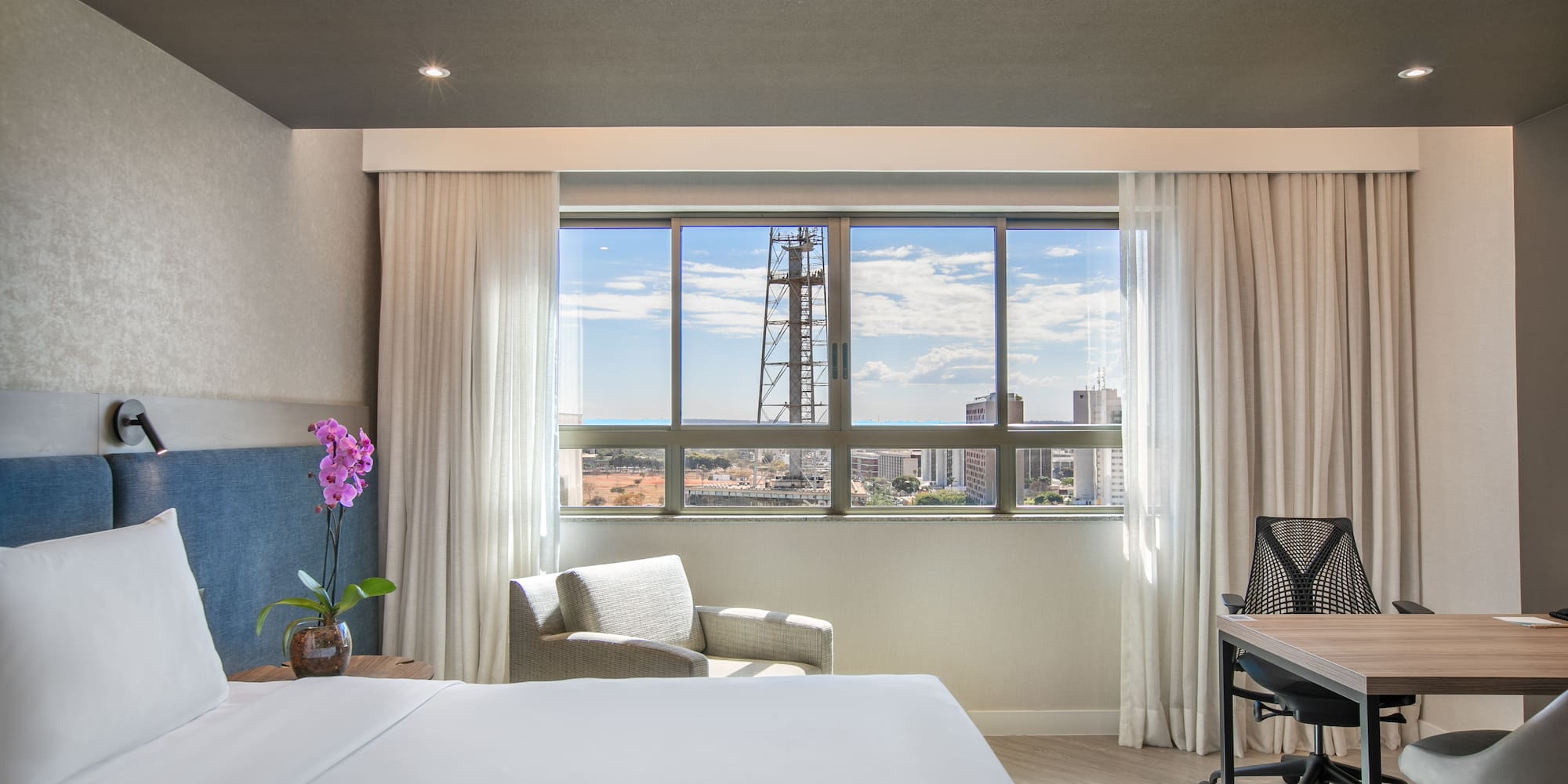 Modern hotel room with city view, white bed, armchair, and work desk.