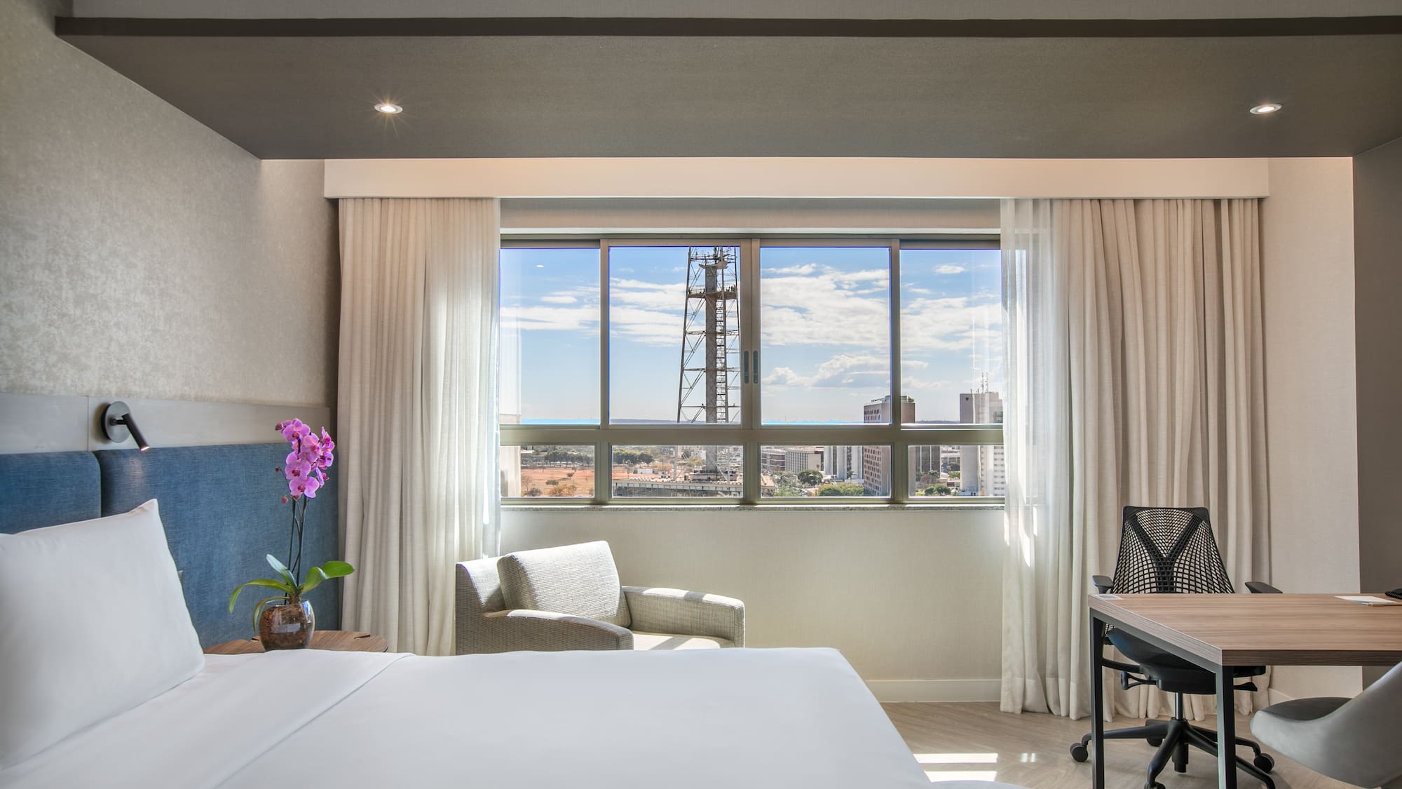 Modern hotel room with city view, white bed, armchair, and desk.
